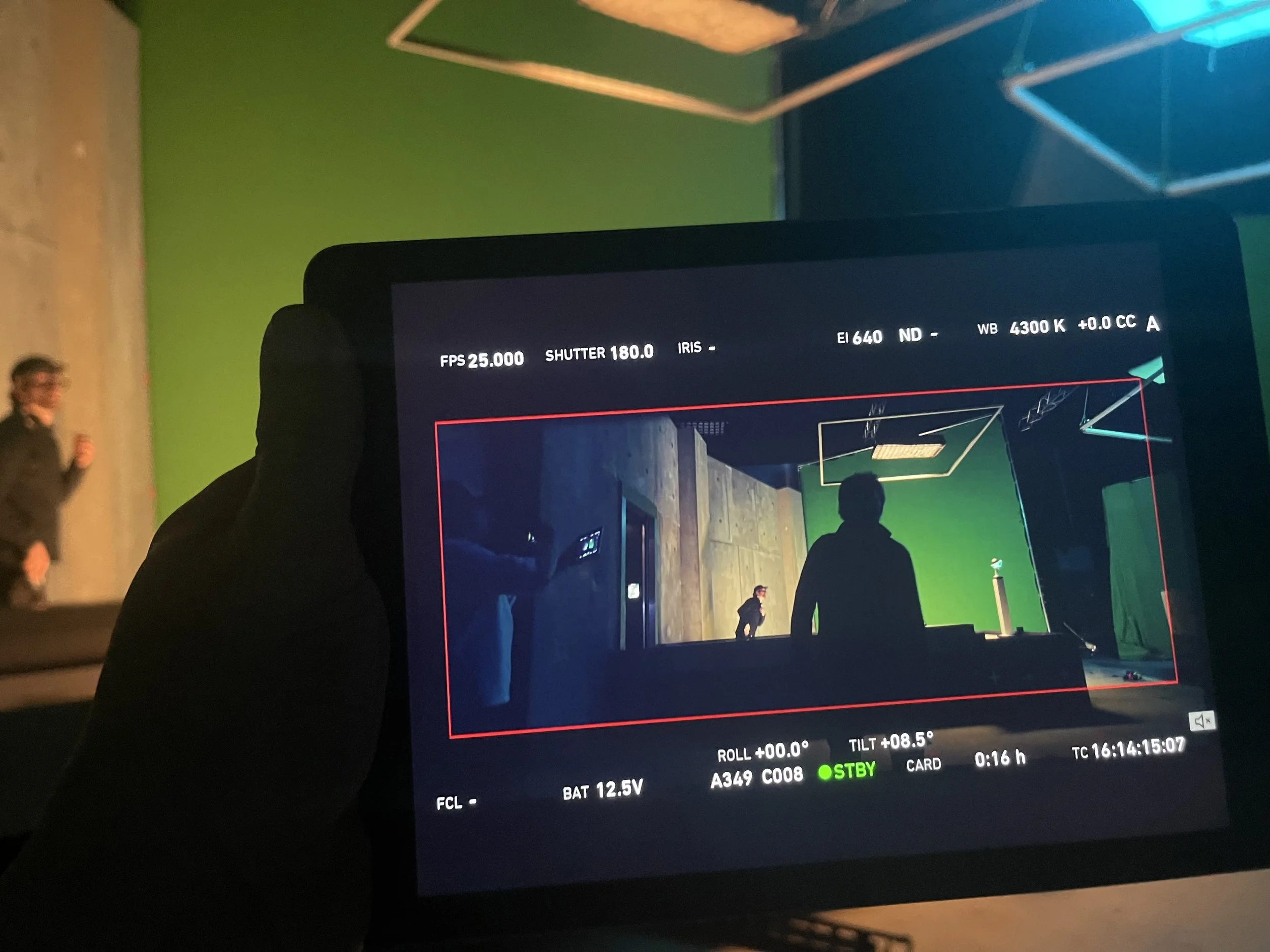 The image shows a digital camera monitor displaying a photograph in a studio setting. The photo on the monitor features a green screen background and people in front of it. Camera settings such as FPS, shutter speed, ISO, white balance, and battery s