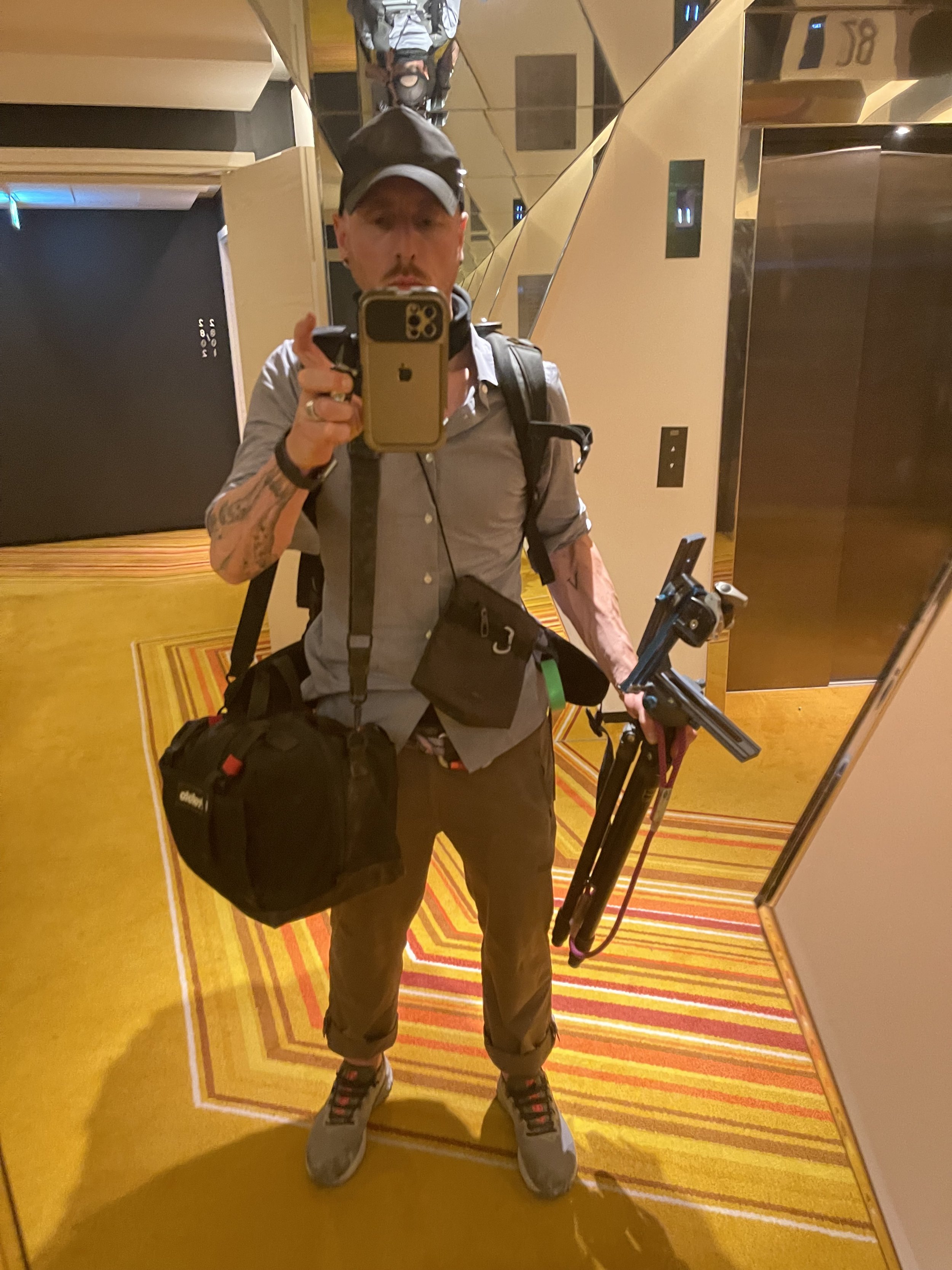 A person taking a mirror selfie in an elevator. They are wearing a gray shirt, brown pants, a black cap, and have tattoos on their left arm. They are carrying a large black backpack and holding a tripod with a camera mounted on it.