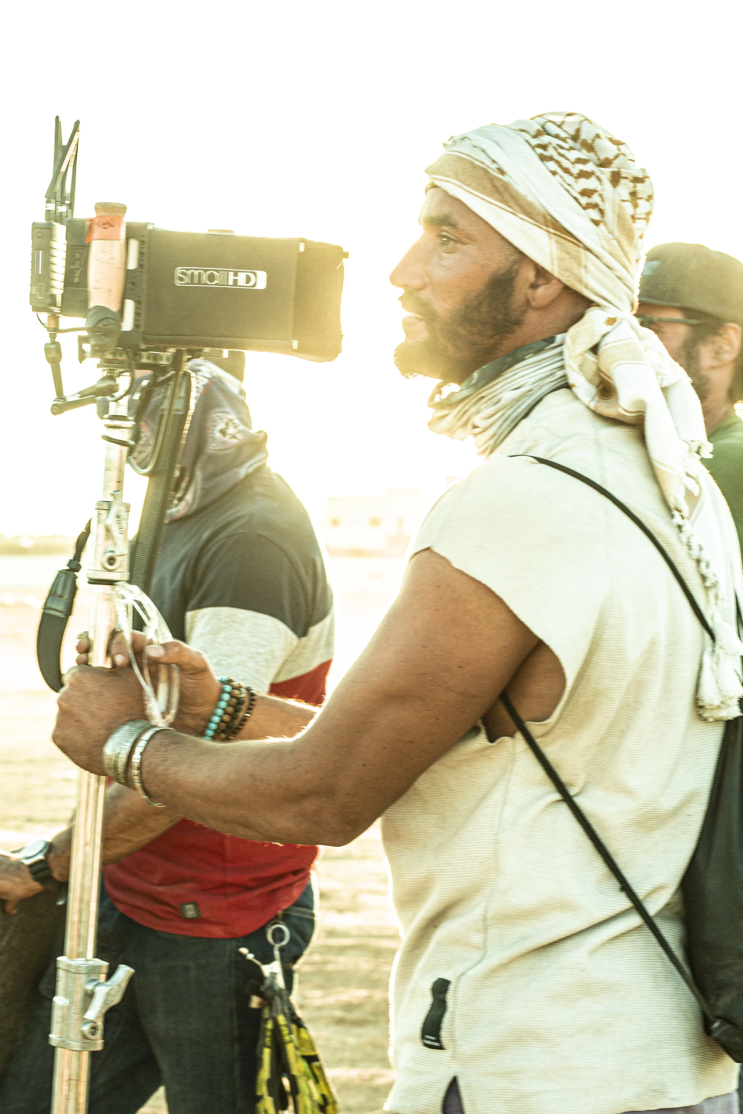 People filming outdoors during sunset, with Bilall Fallah in the foreground wearing a headscarf and a sleeveless shirt, operating a professional camera on a tripod. Photo taken by Kris Janssens on the set of the feature film Rebel.