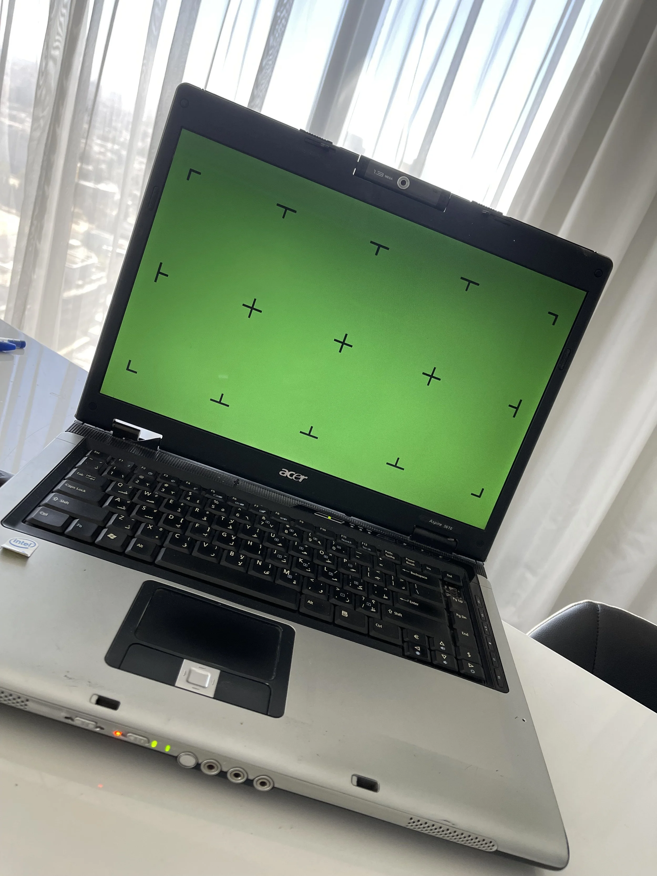 A laptop with a green screen displaying crosshair markings, sitting on a white surface with a computer mouse underneath, in front of sheer white curtains allowing diffused sunlight.