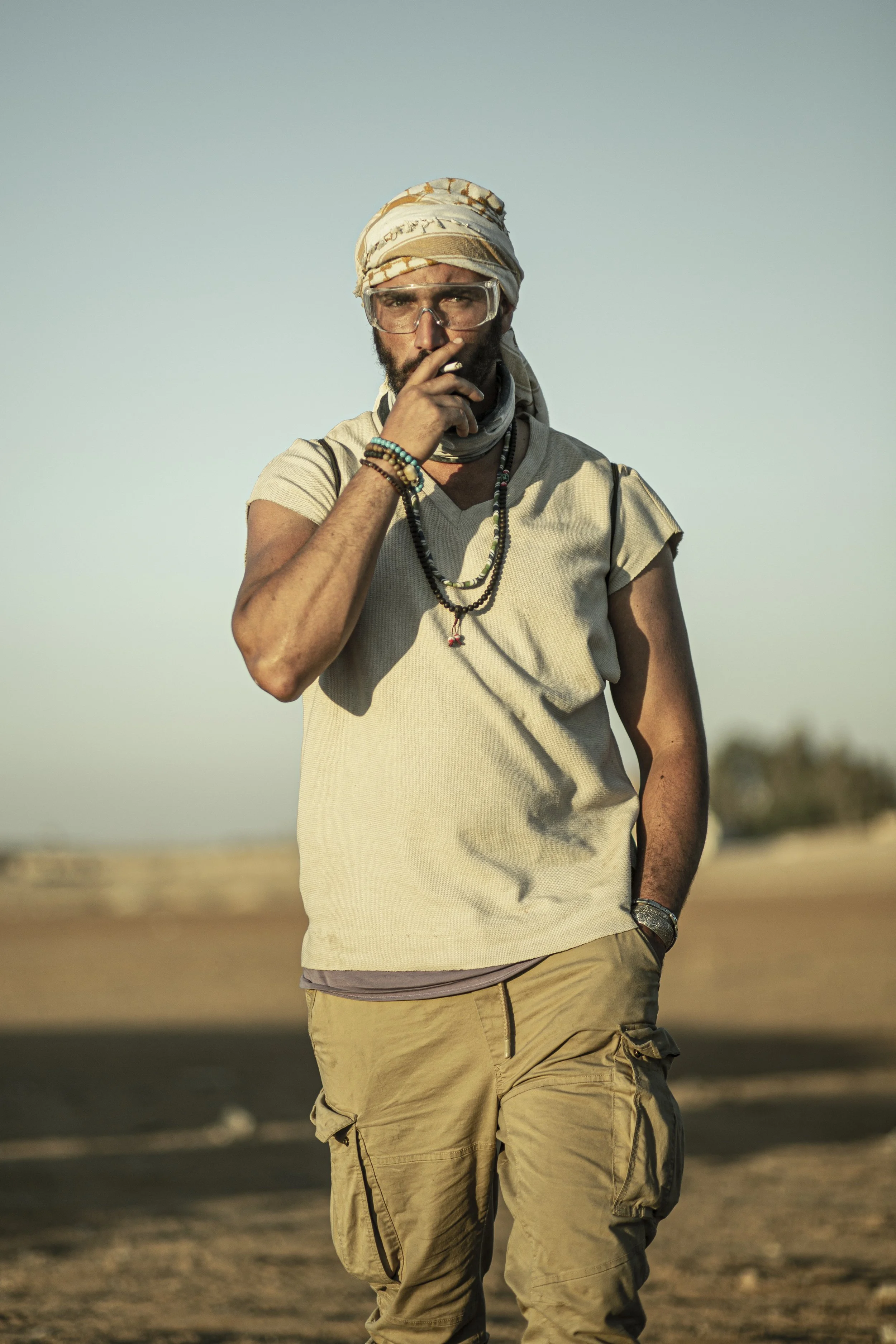 Bilall Fallah with a beard wearing beige cargo pants and a beige t-shirt, accessorized with bracelets and necklaces, standing outdoors with a clear sky in the background.  Photo taken by Kris Janssens on the set of the feature film Rebel.