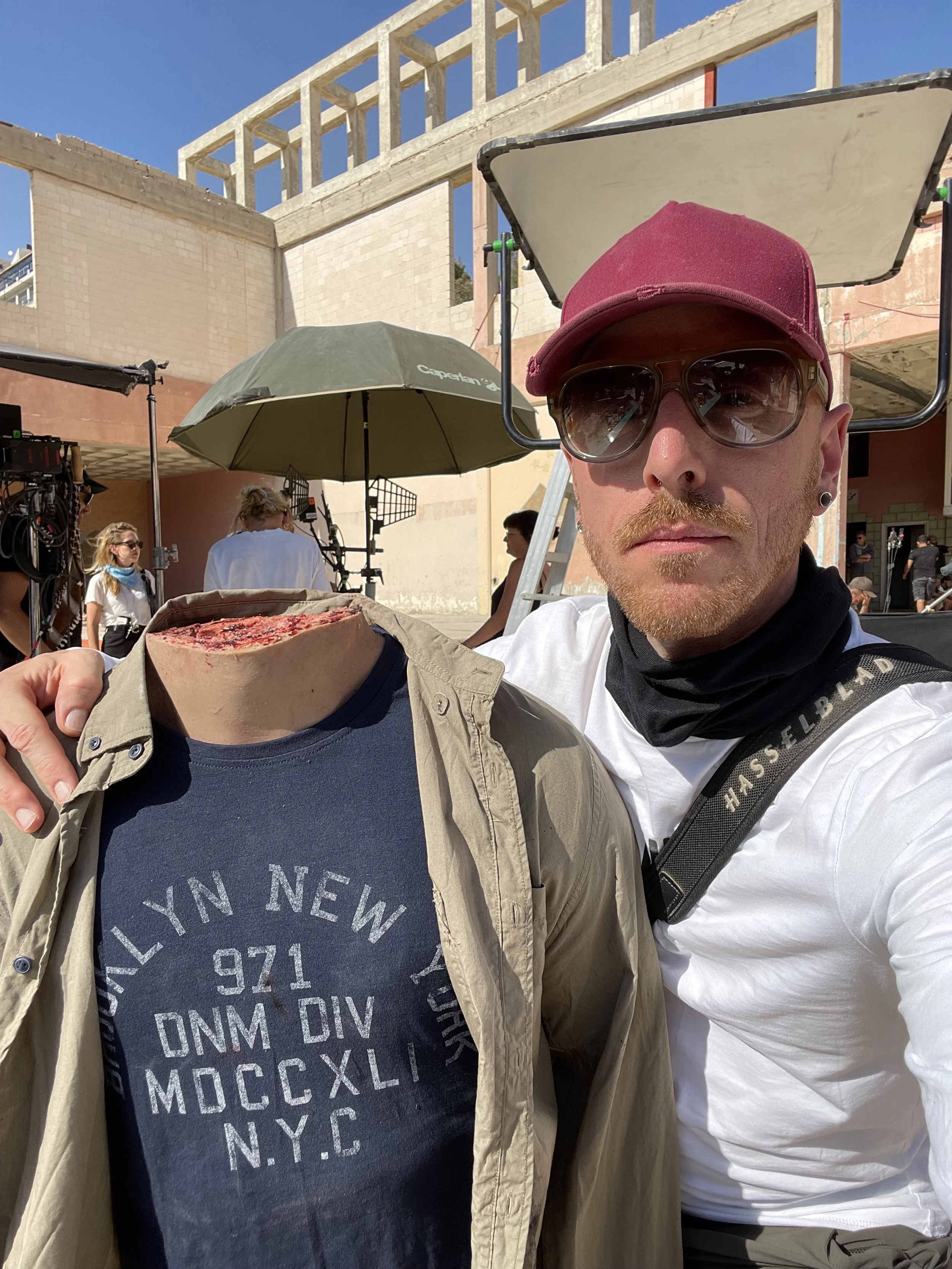 A man wearing a red cap, sunglasses, a white shirt, and a black neck gaiter taking a selfie at an outdoor film or photo shoot. He is holding a beige jacket over a navy blue T-shirt with printed text. In the background, there are film equipment, a wom