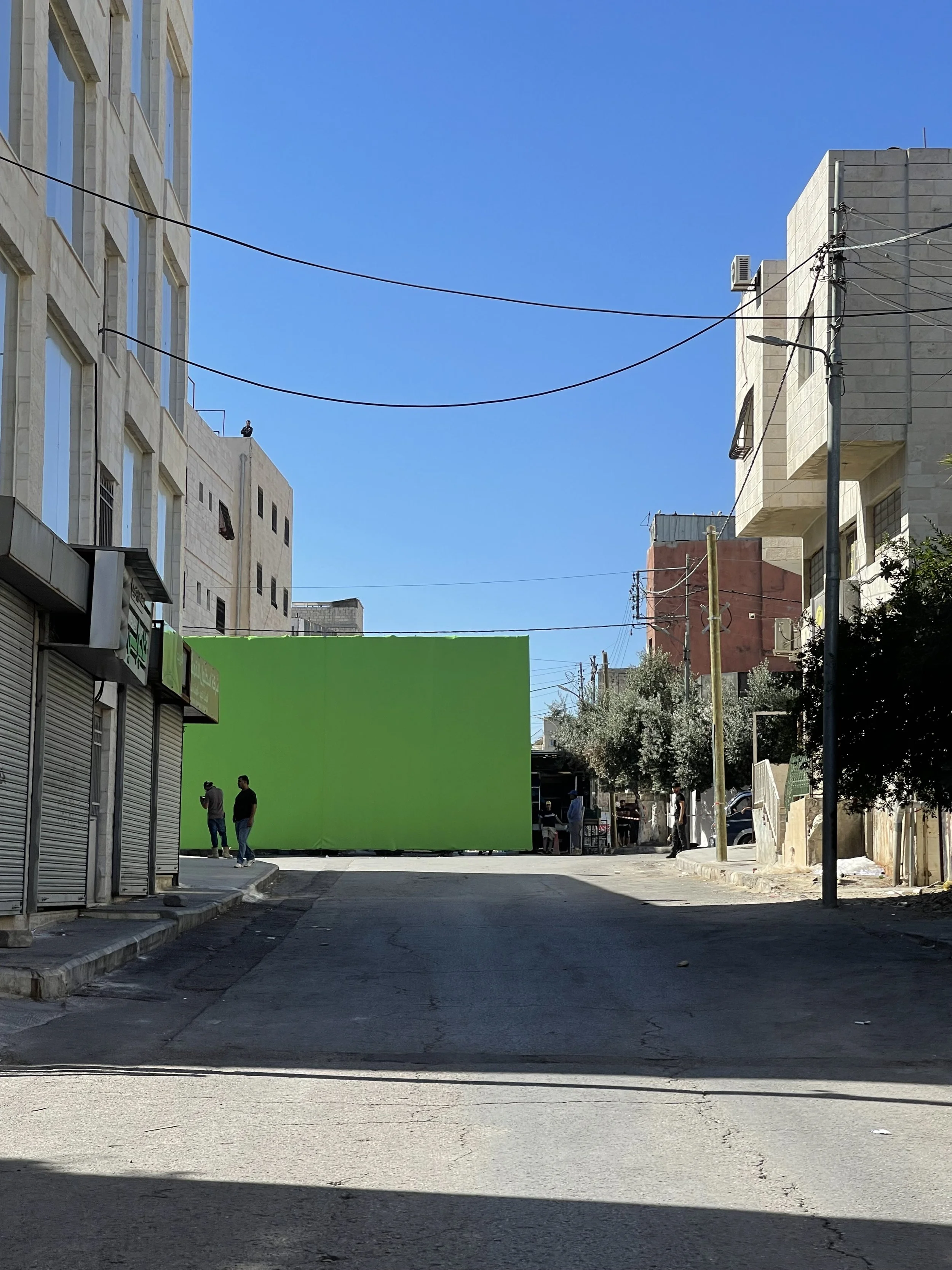 A city street scene with buildings, electrical wires, and a group of people standing near a green screen backdrop.