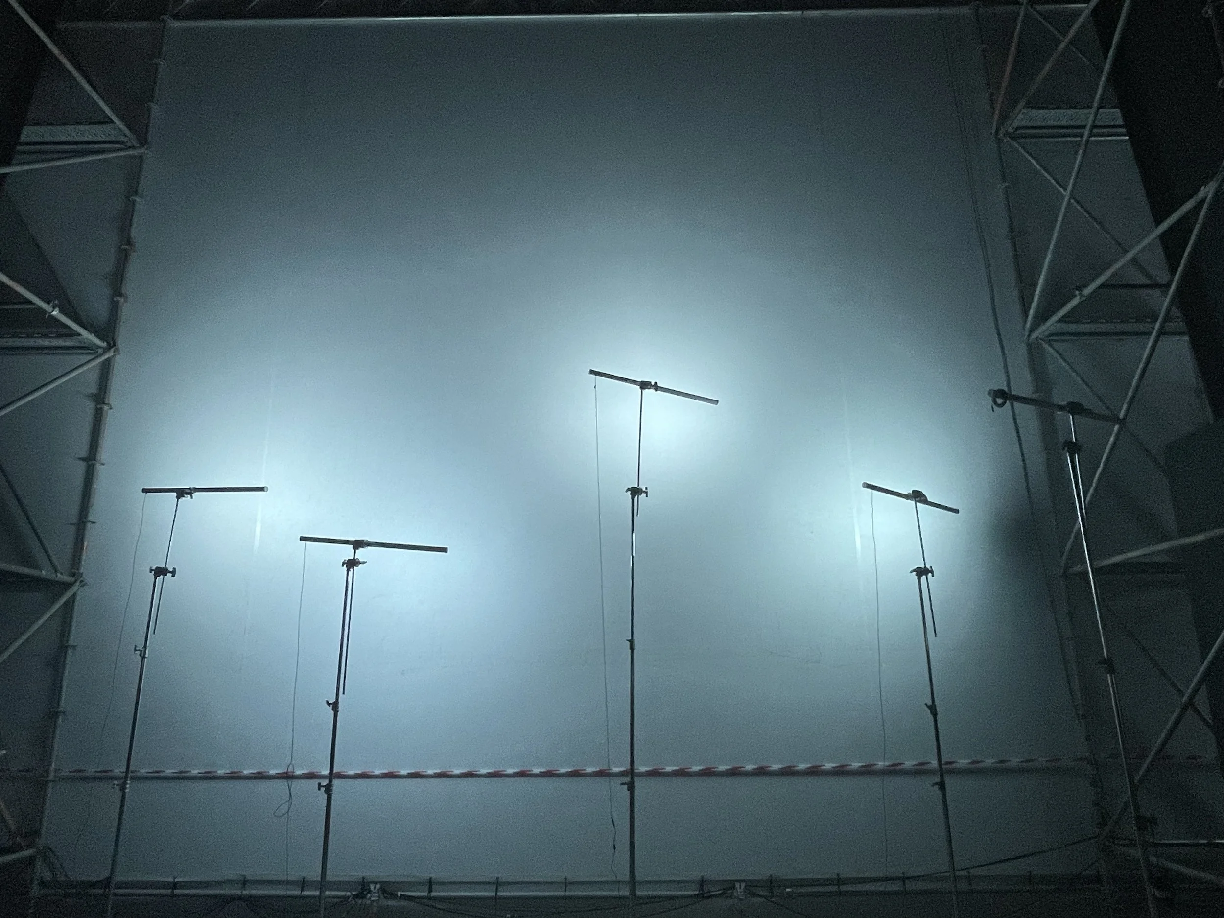 The image depicts a construction or studio setup with a large, bright light source shining upwards on a high ceiling. Metal scaffolding frames surround the scene on both sides, with lights or equipment attached to the scaffolding. The overall atmosph