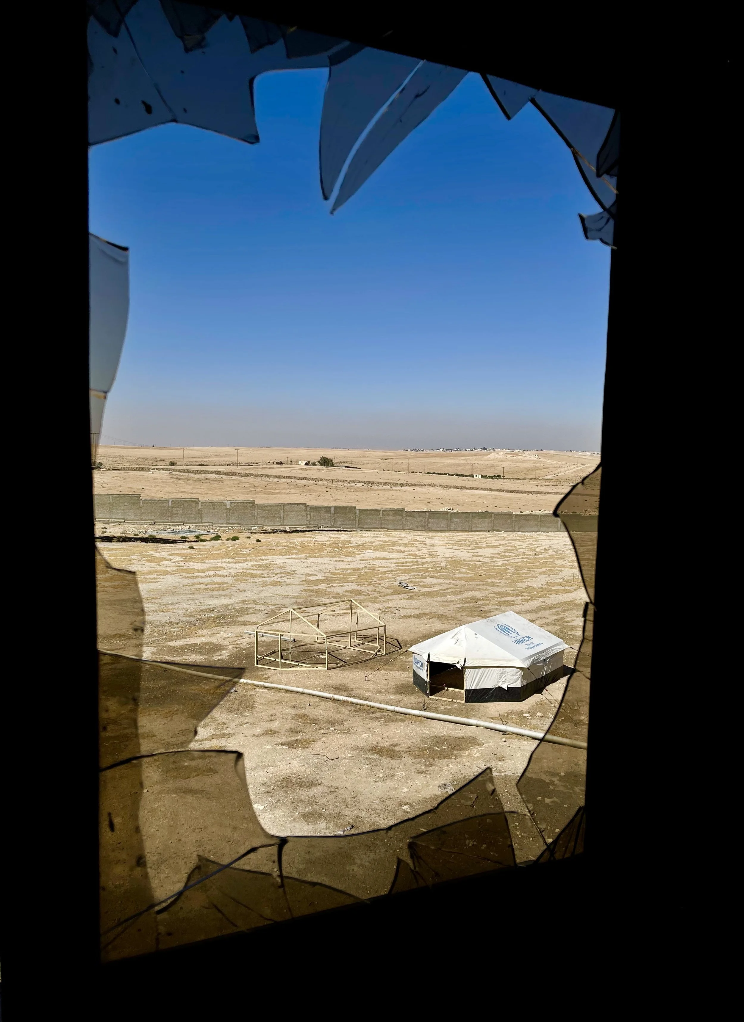 Photo of a desert landscape seen through a broken window with shattered glass and a tent on the ground.