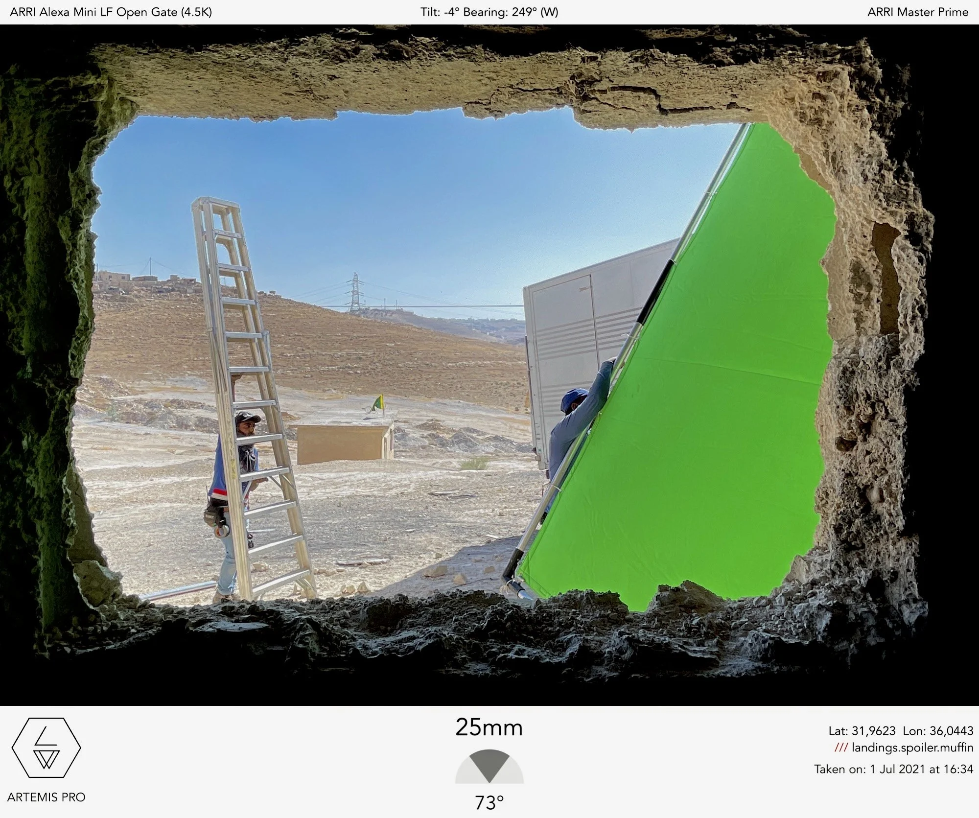 The image shows a view through a rocky opening at the sky, a ladder, and some equipment, with a green screen used for visual effects, and a person working with some equipment in an outdoor area with hills, utility poles, and a white building.