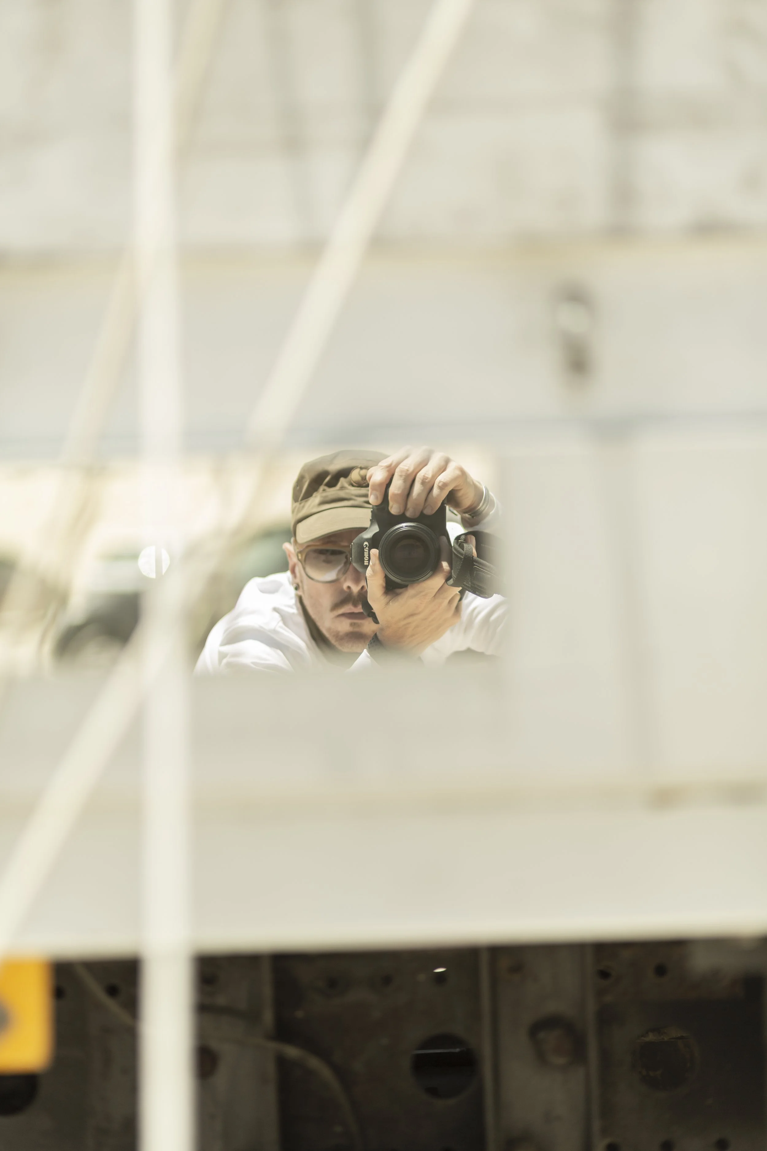 A person in a hat and glasses taking a photo of their reflection with a camera, seen through a reflective surface or mirror, with an industrial or architectural background.