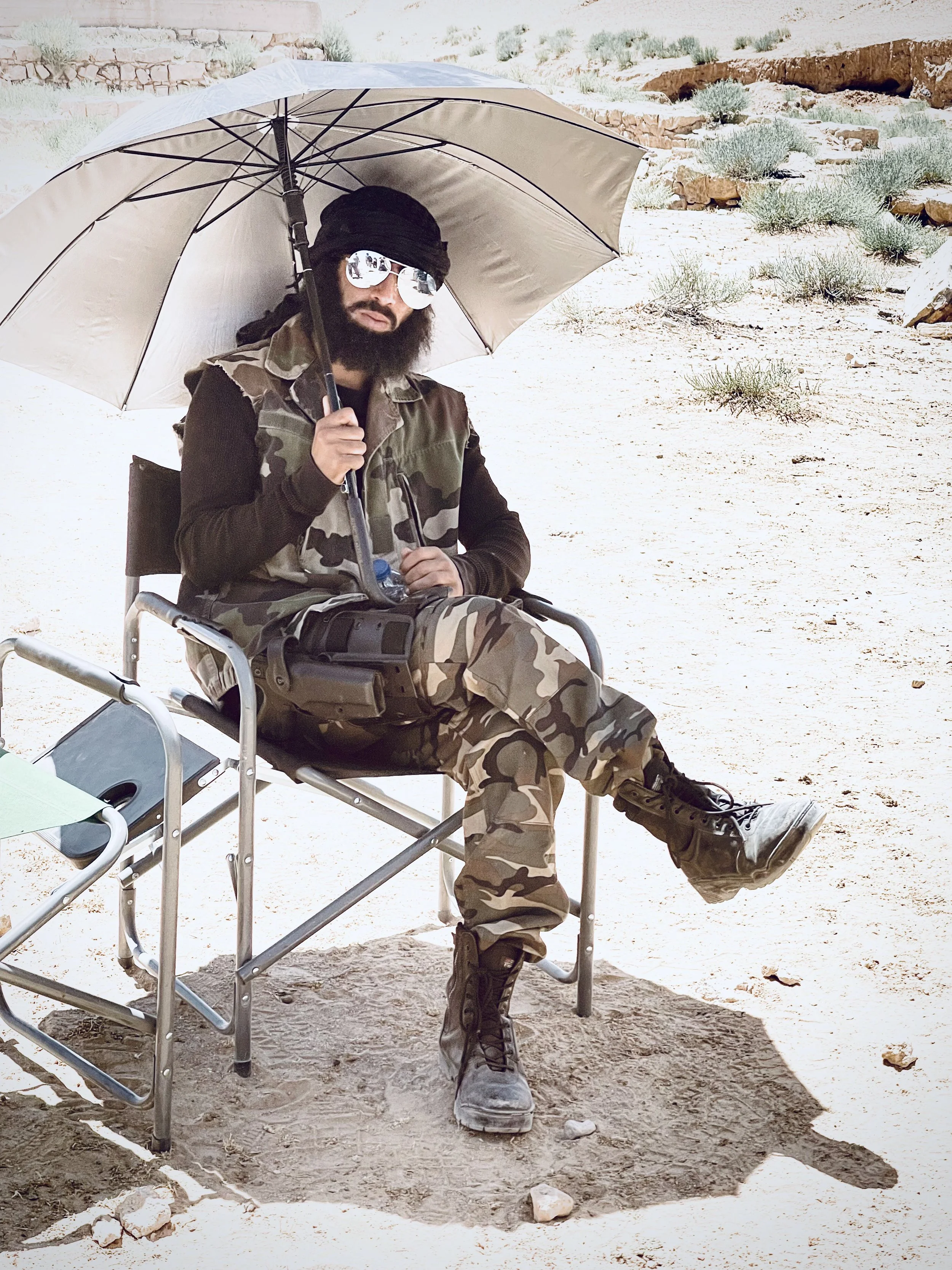 A man with a beard, sunglasses, and black head covering sitting on a camping chair, holding an umbrella in a desert landscape with sparse bushes and rocks.