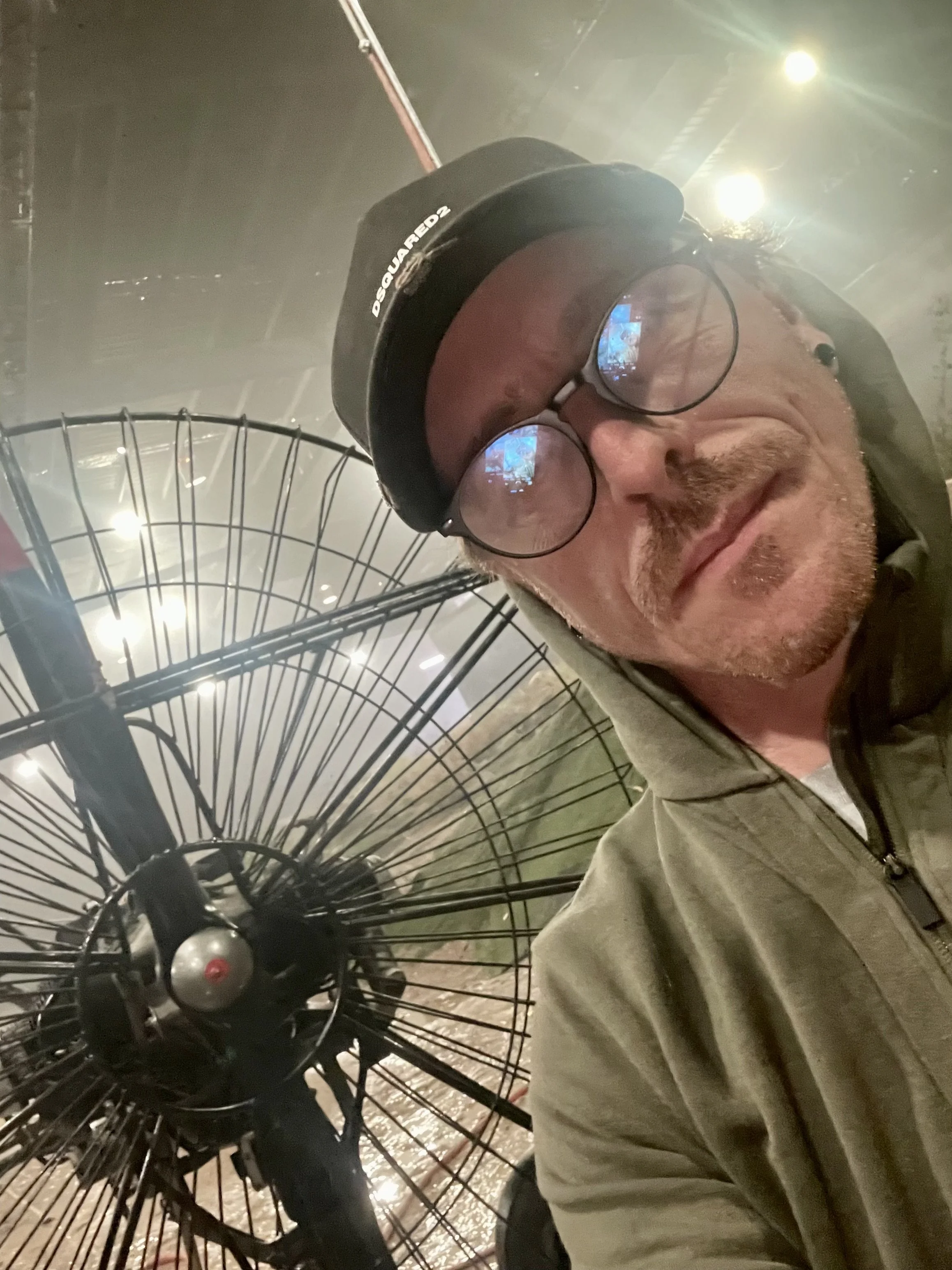 A man with glasses and a beard wearing a black helmet and tan jacket is taking a selfie next to a vintage bicycle with large front and small rear wheels in an indoor setting with bright overhead lights.