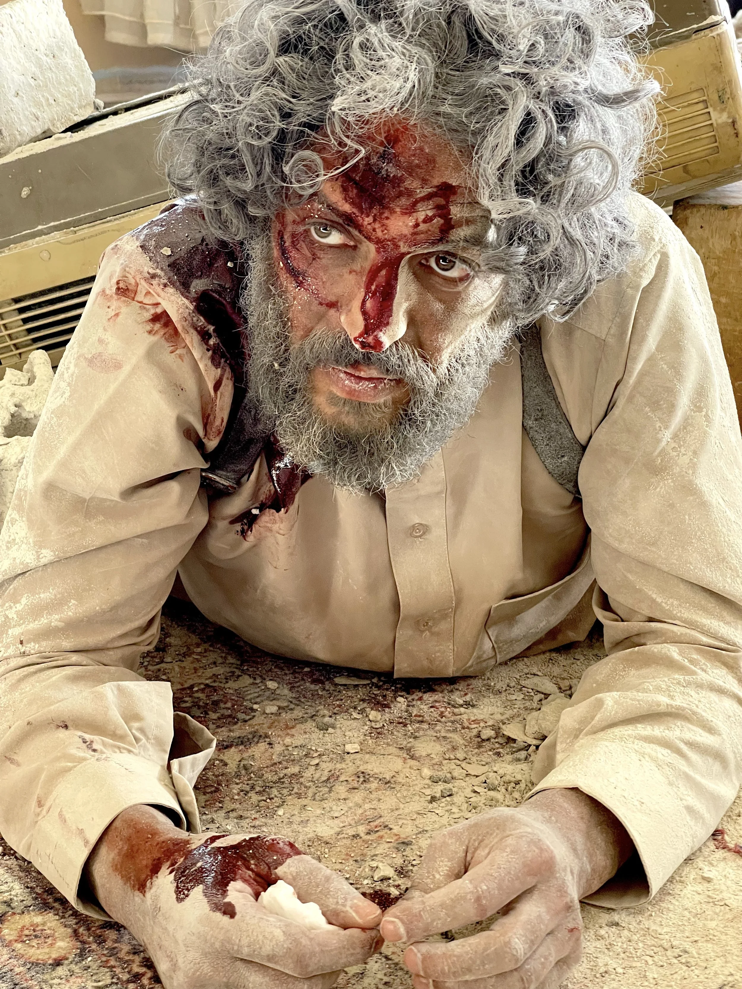 A man with a beard and curly gray hair, dressed in a beige shirt, is sitting on the ground with a serious expression. His face and arm appear to have fake wounds with fake blood, and he looks directly at the camera. The setting appears to be a dusty,