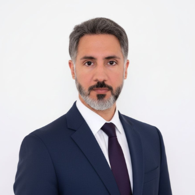 Headshot of a man with gray hair, beard, wearing a navy blue suit, white shirt, and dark tie, against a plain white background.