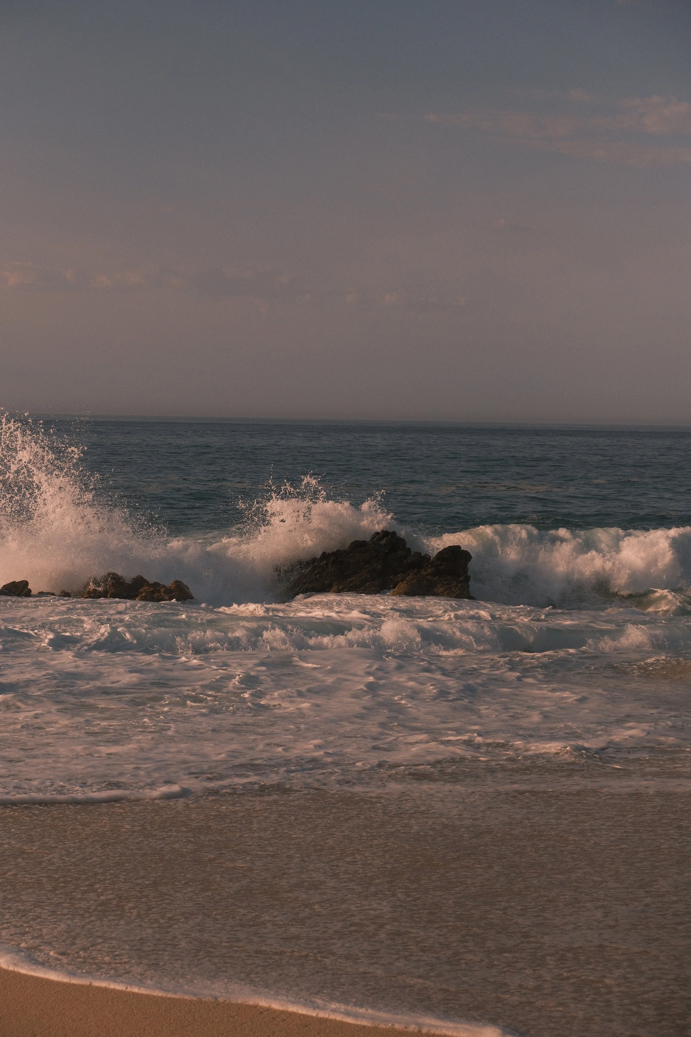 Waves crashing onto rocks on the beach at sunset.