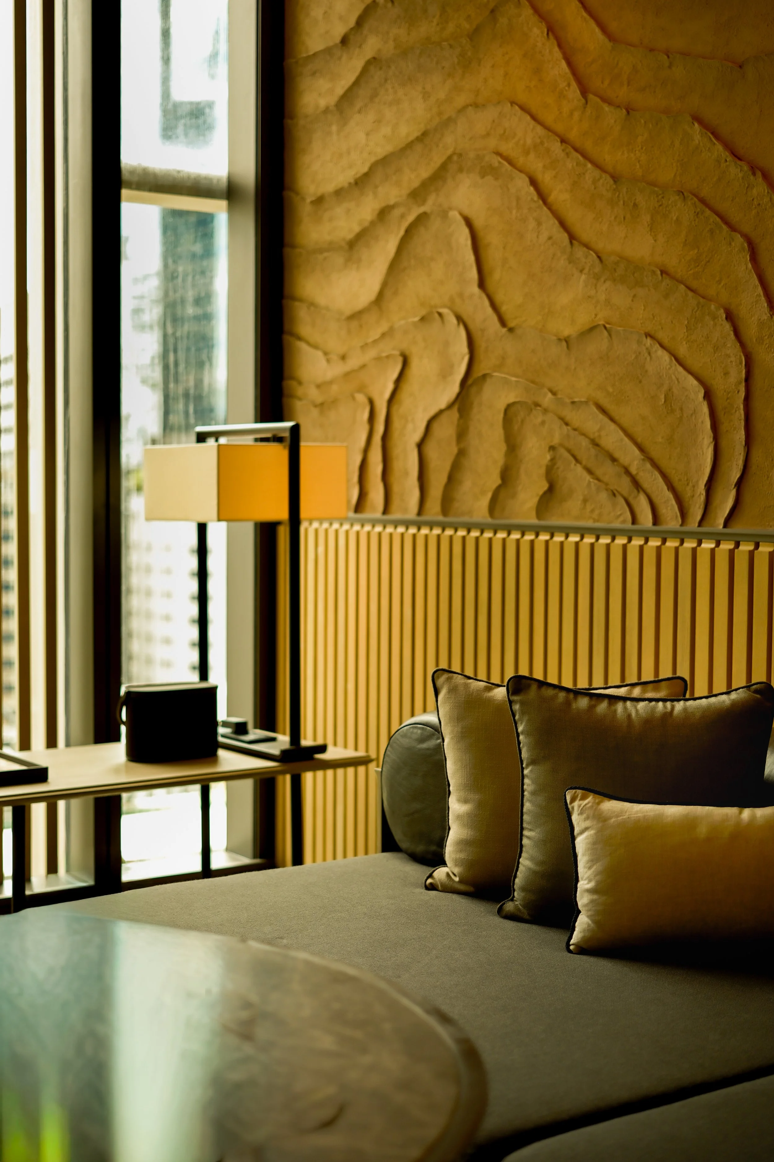 A cozy living room with beige pillows on a soft sofa, a wooden side table with a black lamp, and a large window with vertical blinds letting in natural light. A textured wall with abstract patterns is in the background.