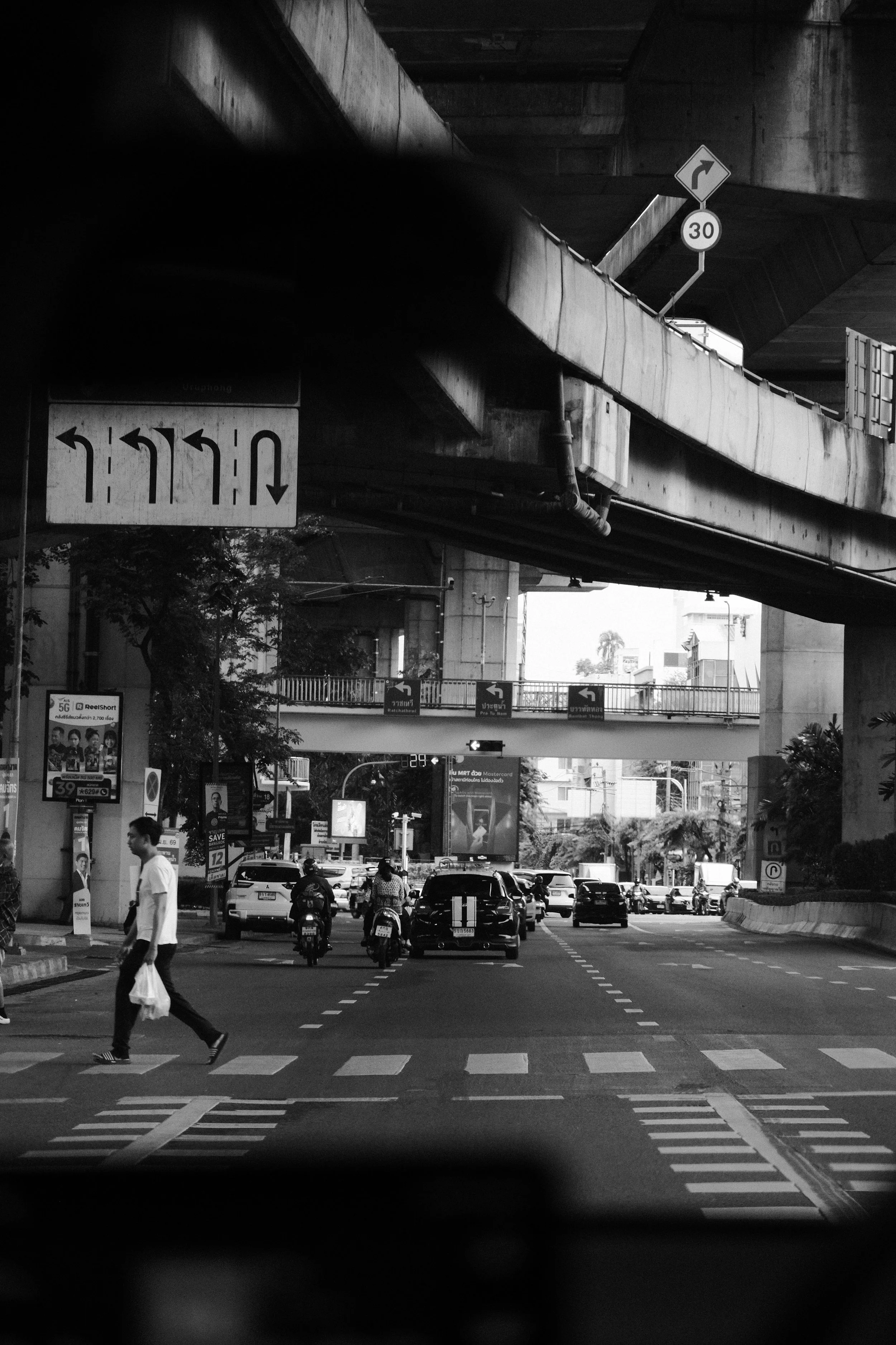 A busy city street scene captured from under a bridge with multiple traffic signs overhead, including a right turn and speed limit signs. Several cars and motorcycles are on the road, and a pedestrian is crossing at a crosswalk. The scene is in black