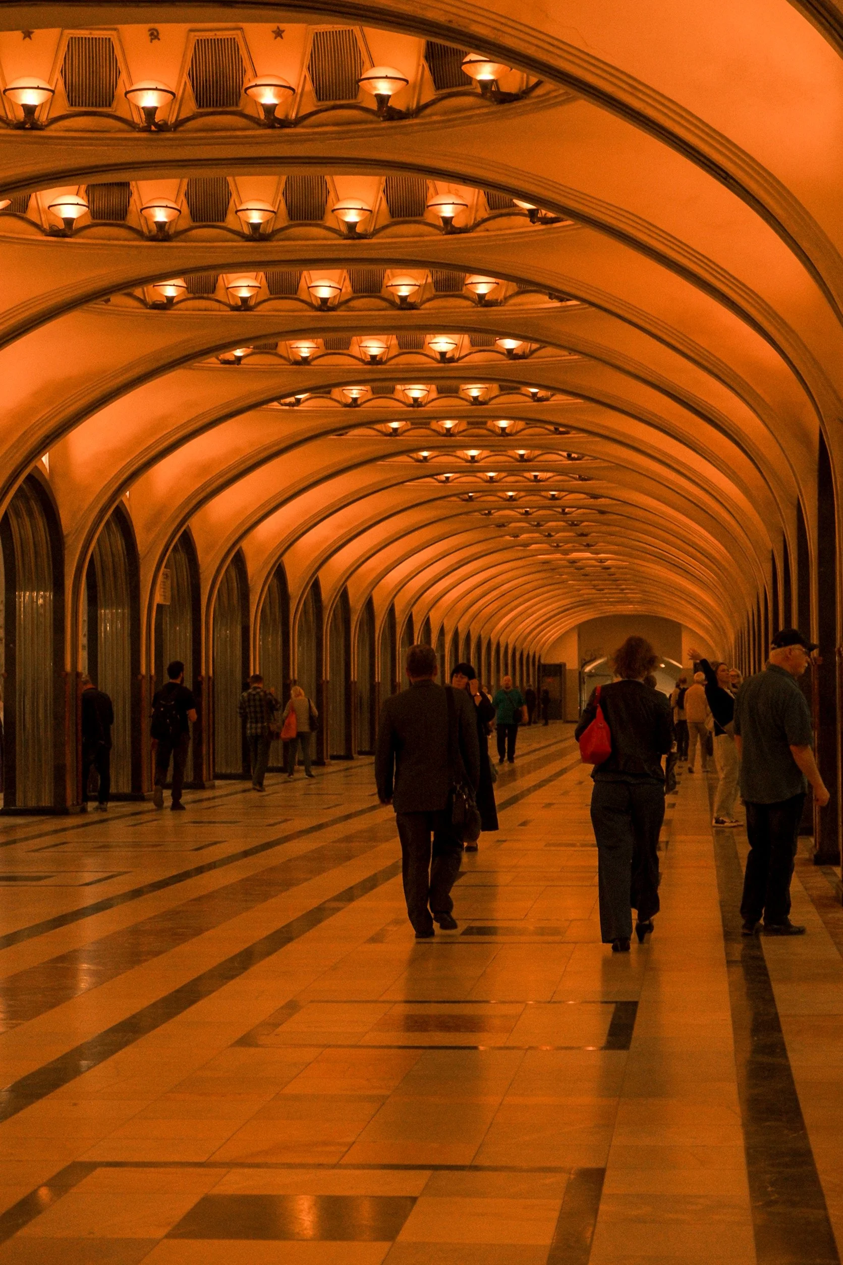 People walking through an arched hallway with warm lighting and reflective brown tile flooring.