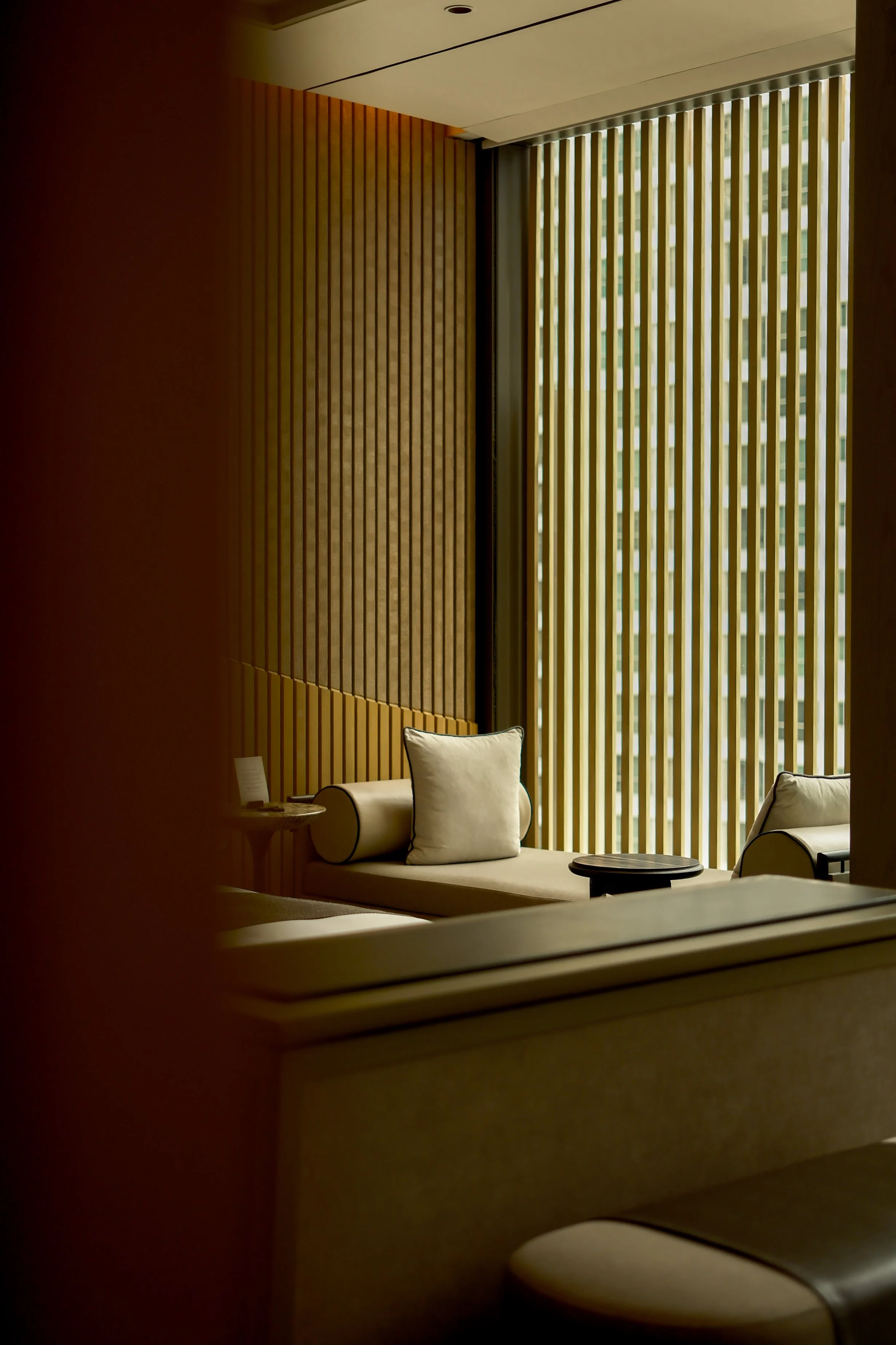 Interior of a modern lounge with beige chairs, a small round table, and wooden wall panels, next to a large window with vertical blinds.