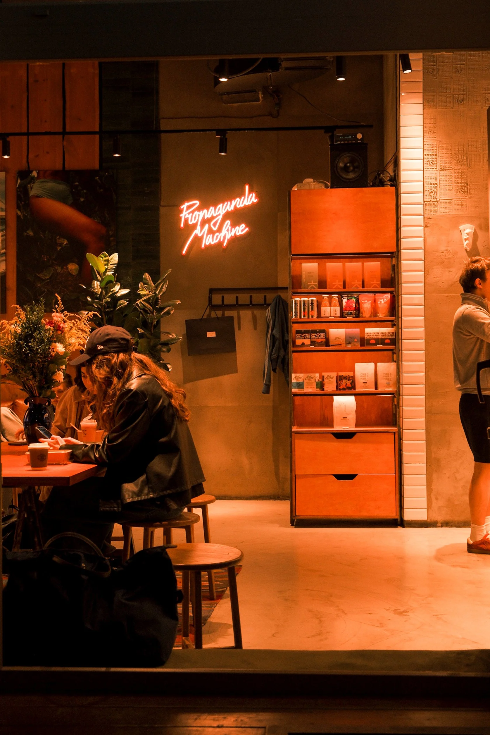 A busy indoor cafe scene with people sitting at tables, plants, and a neon sign reading 'Fuqngonda Machine'. The background includes shelves with books or magazines and a person standing near the shelf. Warm lighting creates a cozy atmosphere.
