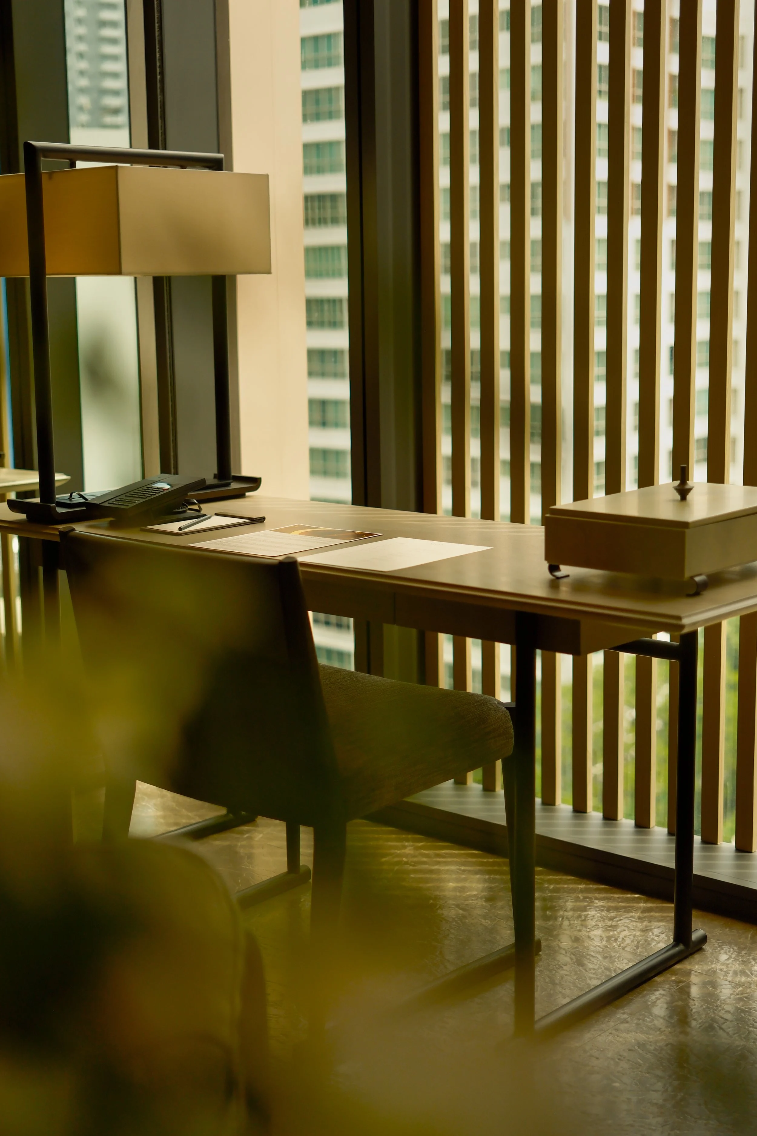 An office desk near a large window with vertical wooden blinds, a lamp, a telephone, some papers, and a sushi box on the desk. The window shows a view of tall buildings outside.