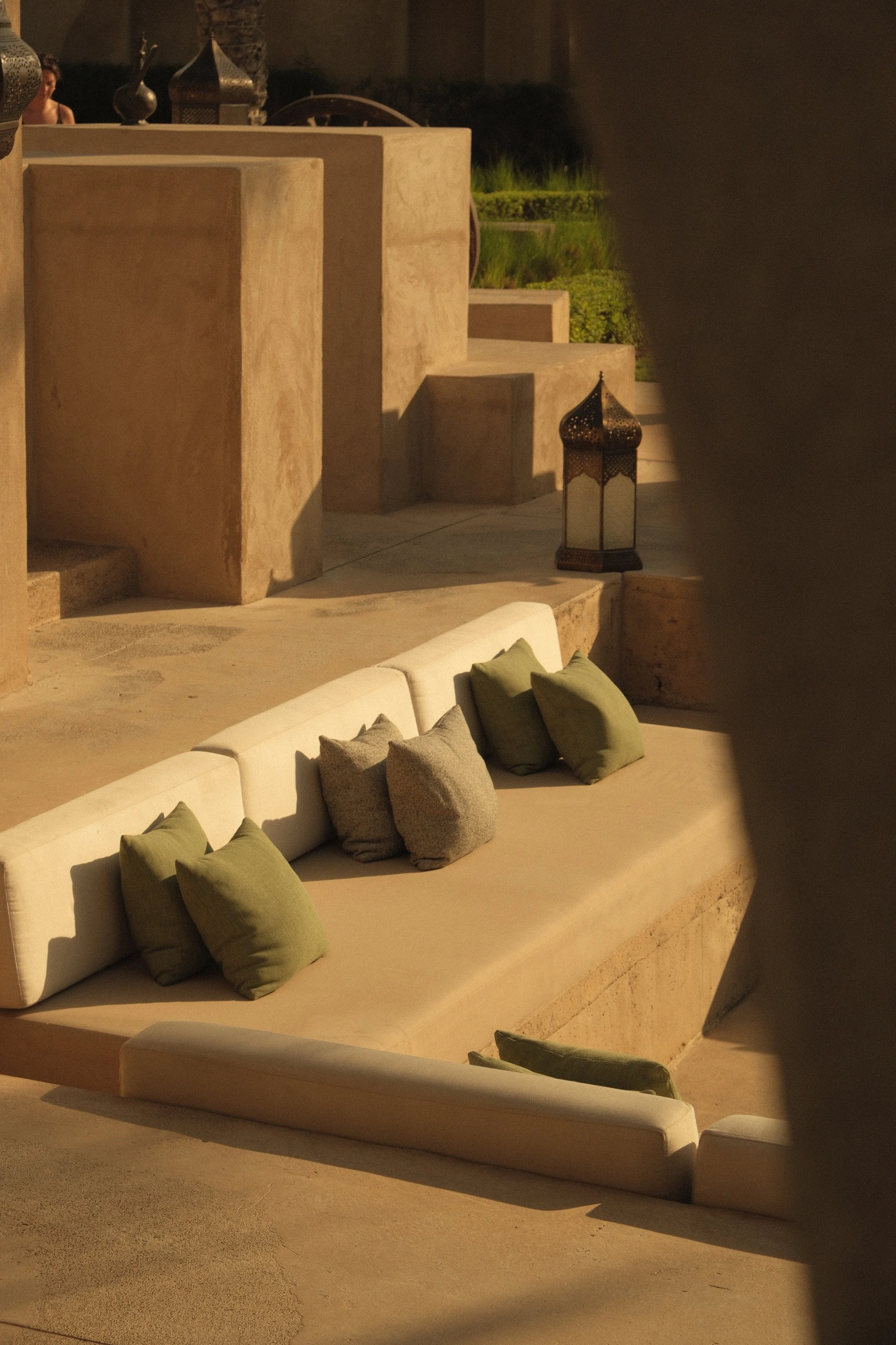 Outdoor seating area with a long beige cushioned bench decorated with green and gray pillows, a traditional lantern, and staircases made of stone leading to a garden with greenery and decorative vases.
