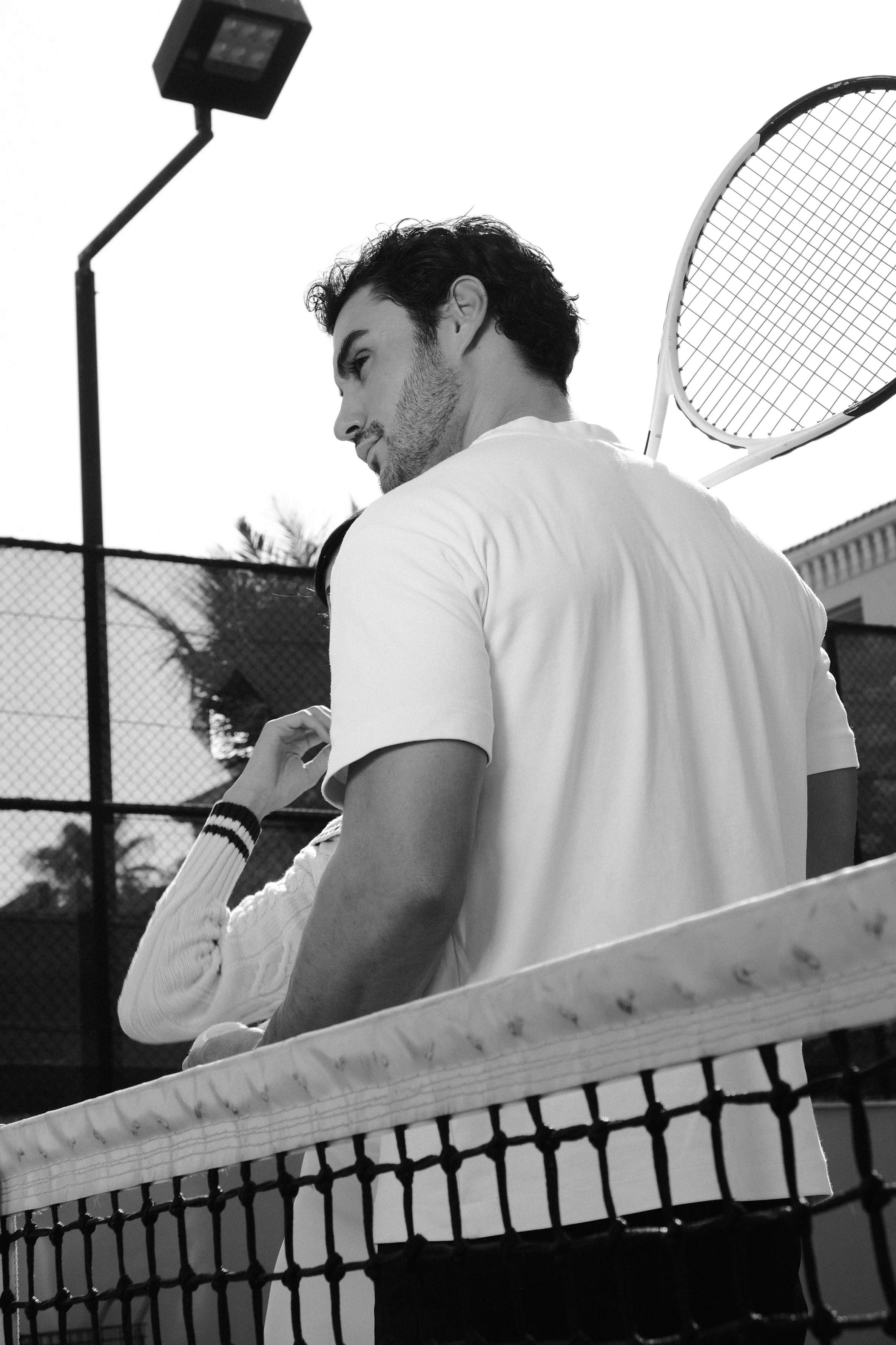 A black-and-white photo of a man with dark, wavy hair and a beard, holding a tennis racket behind him near a tennis court. The image is taken from below, showing part of the court's net in the foreground and a bright sky in the background.