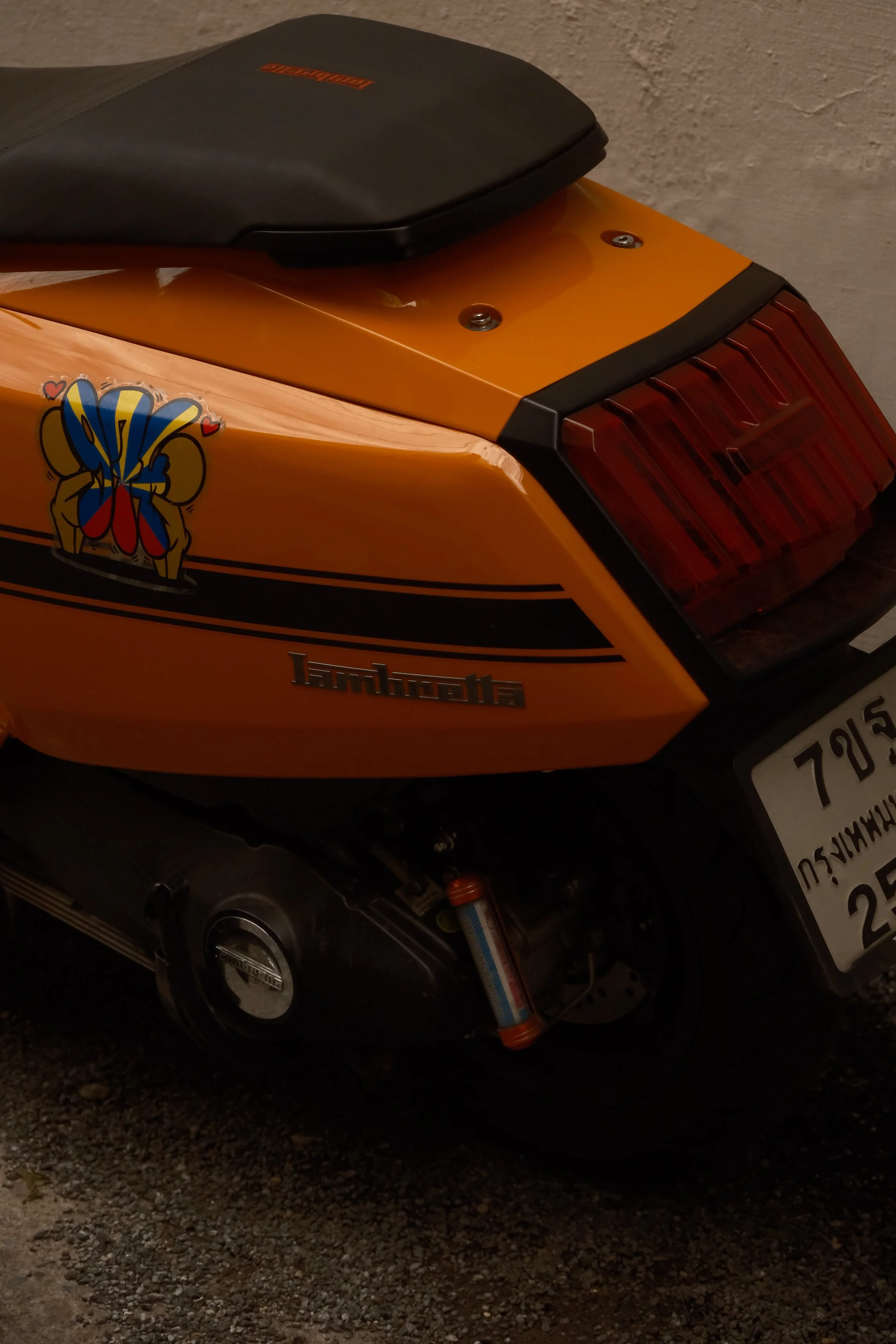 Close-up of a yellow scooter with a black seat, decorated with a colorful sticker of a bee and red hearts, parked on a gravel surface against a textured wall.