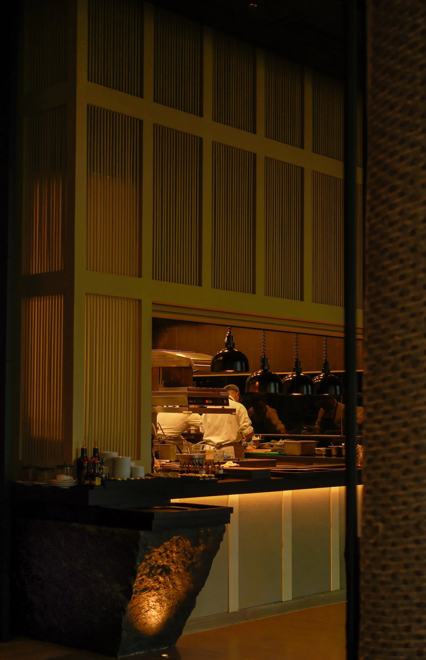 A view into a restaurant kitchen through a window, showing chefs preparing food under black pendant lights, with a decorative wooden partition on the side and a rocky feature below.