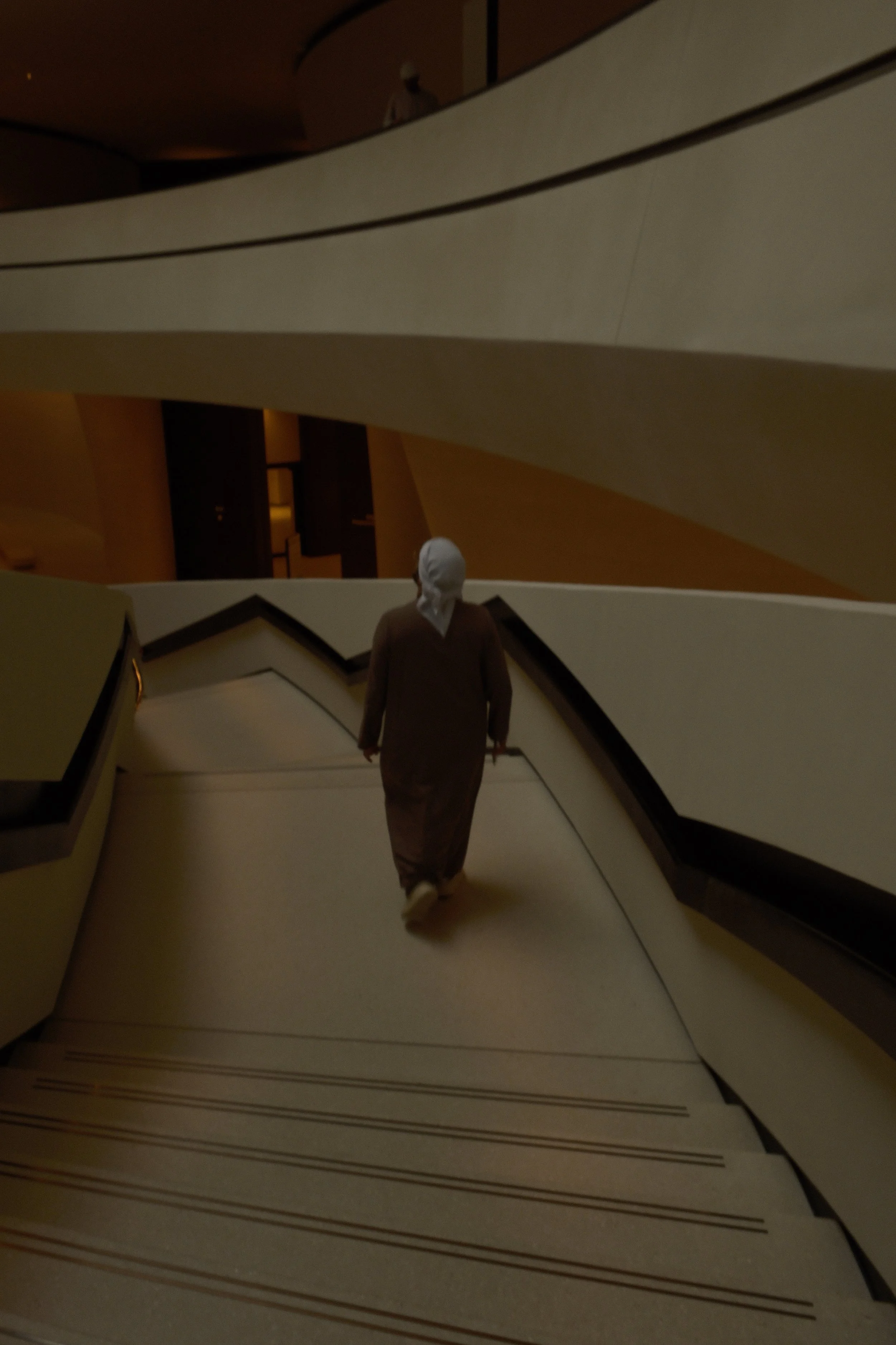 A person wearing a white headscarf and brown clothing walking down an escalator in a modern, dimly-lit building with curved architecture.