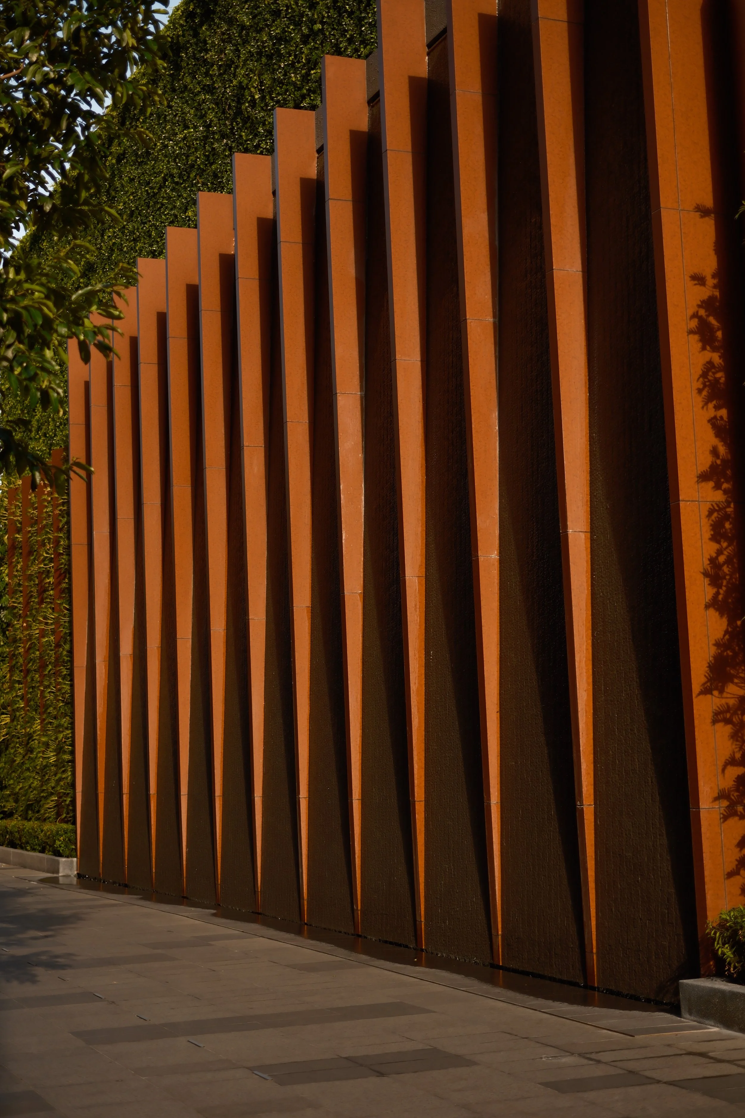 Night view of a modern building facade with vertical, rust-colored panels and a green hedge on top. The ground is paved with large tiles.