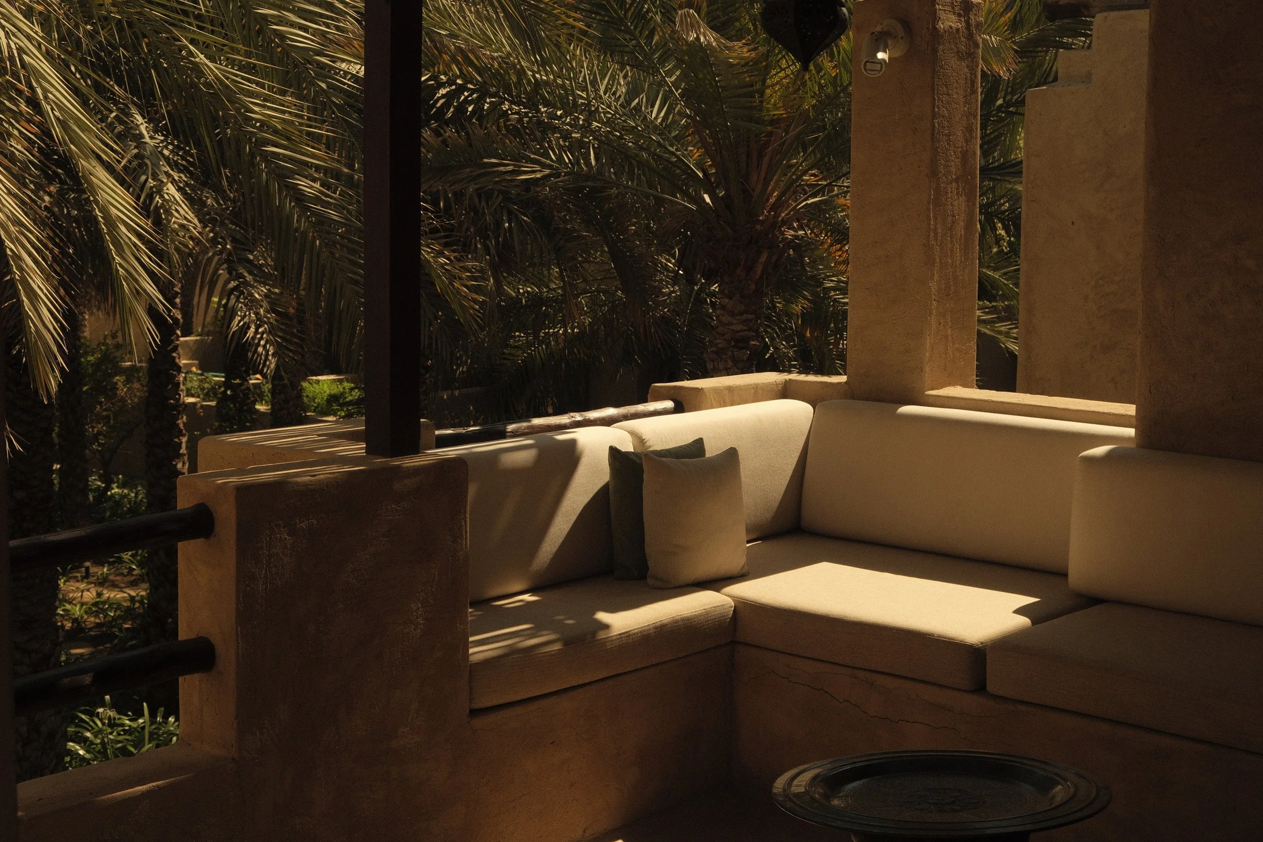 An outdoor patio area with a beige sectional sofa, pillows, and a sound heater, surrounded by lush palm trees and a stone column, during daylight.