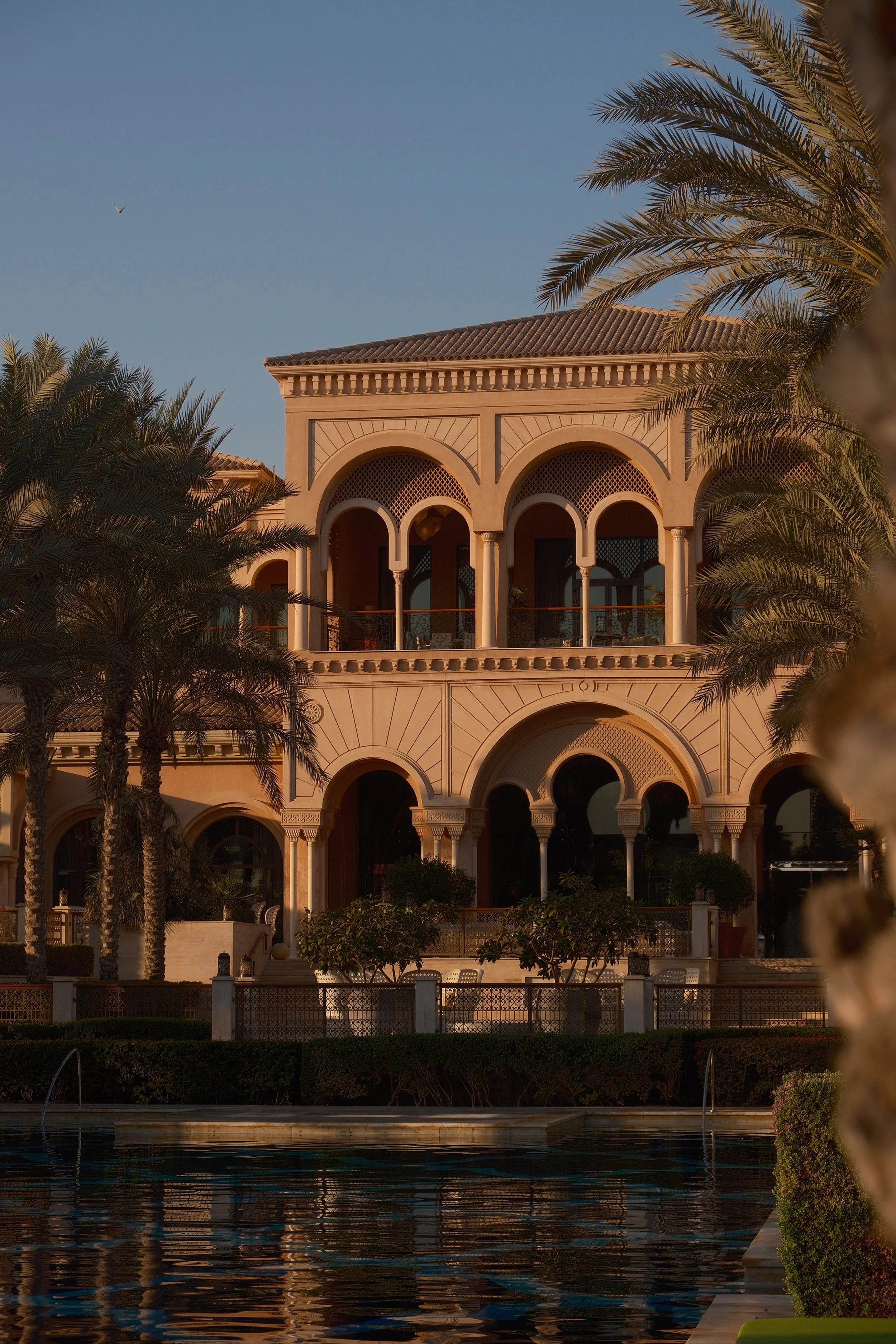 A large Mediterranean-style building with arched windows and a tiled roof, surrounded by palm trees, under a clear blue sky, with a body of water in the foreground reflecting the scene.