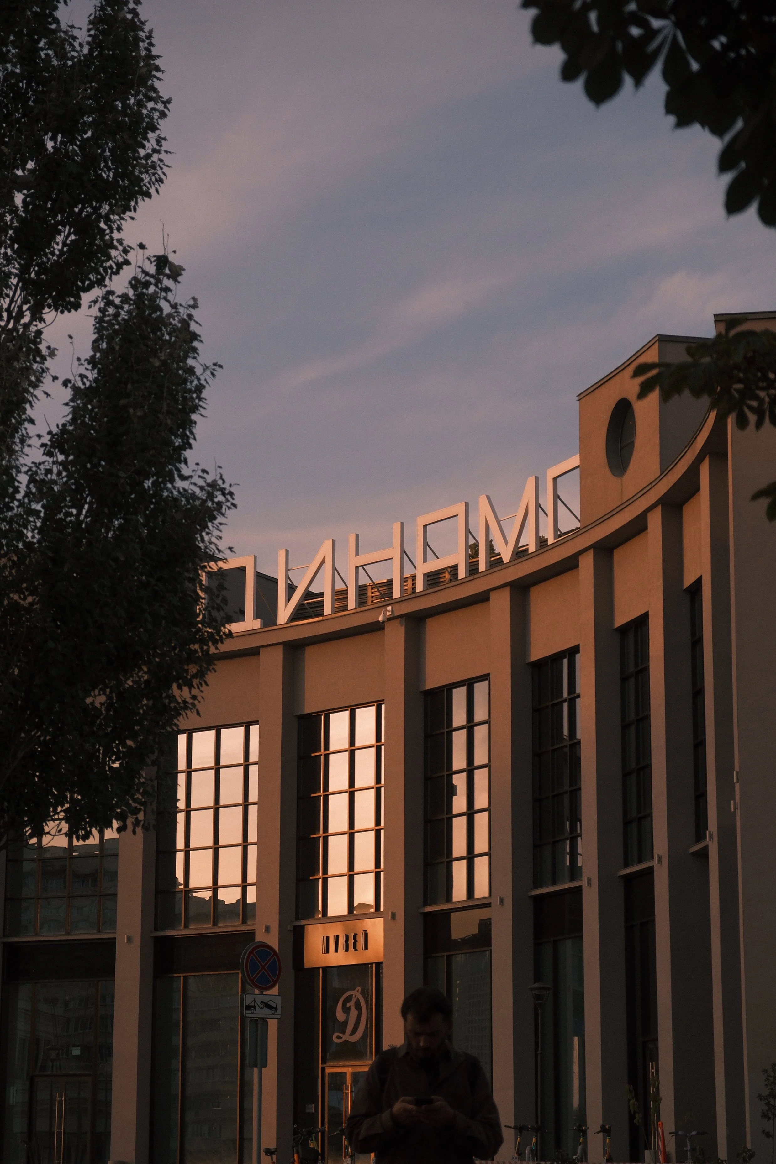 Sunset view of a modern building with large glass windows, trees on the sides, and a person looking at their phone in the foreground.