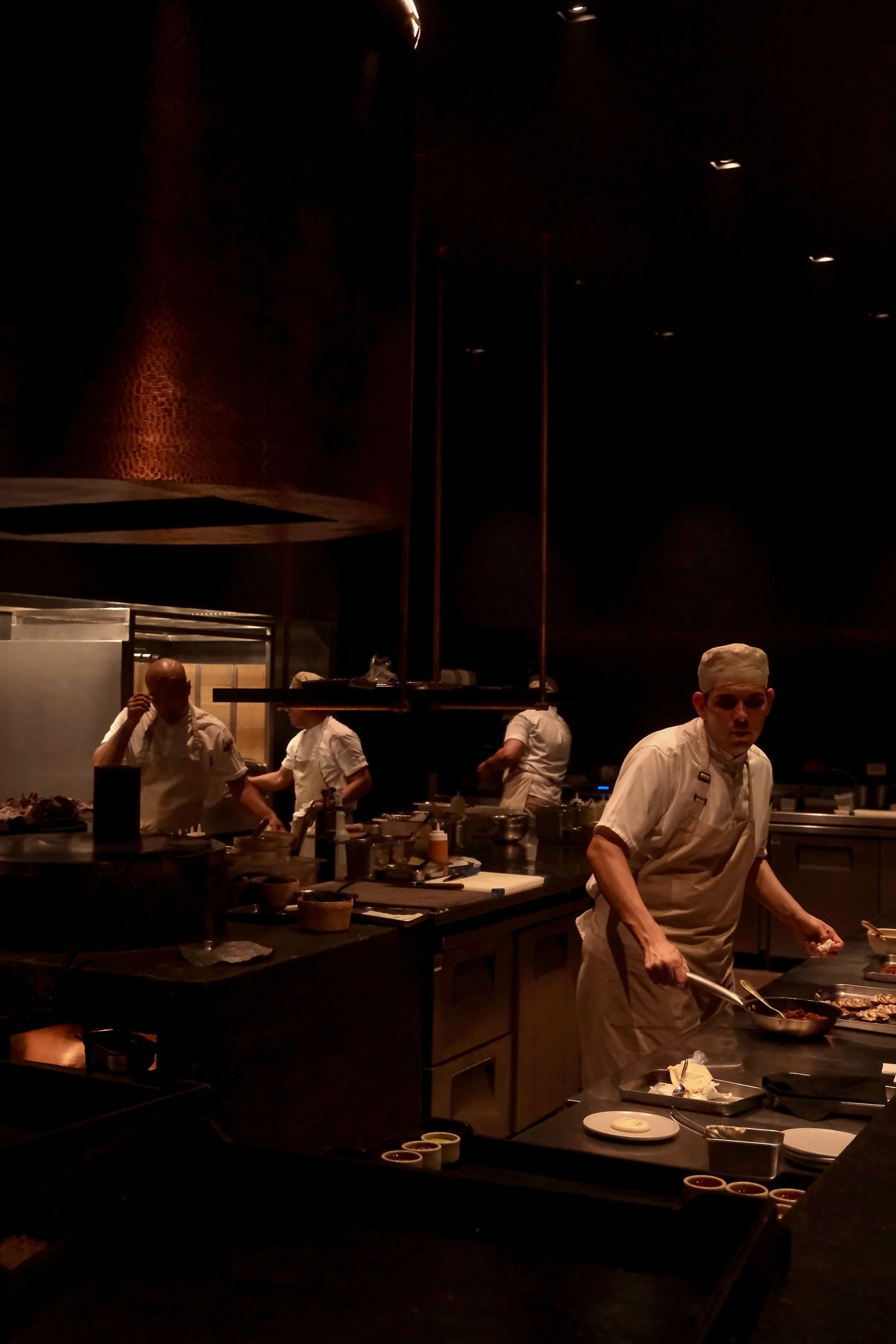 A dimly lit restaurant kitchen with chefs preparing dishes. The scene shows multiple chefs in white uniforms working at various stations, with cooking utensils, plates, and ingredients on the counters.