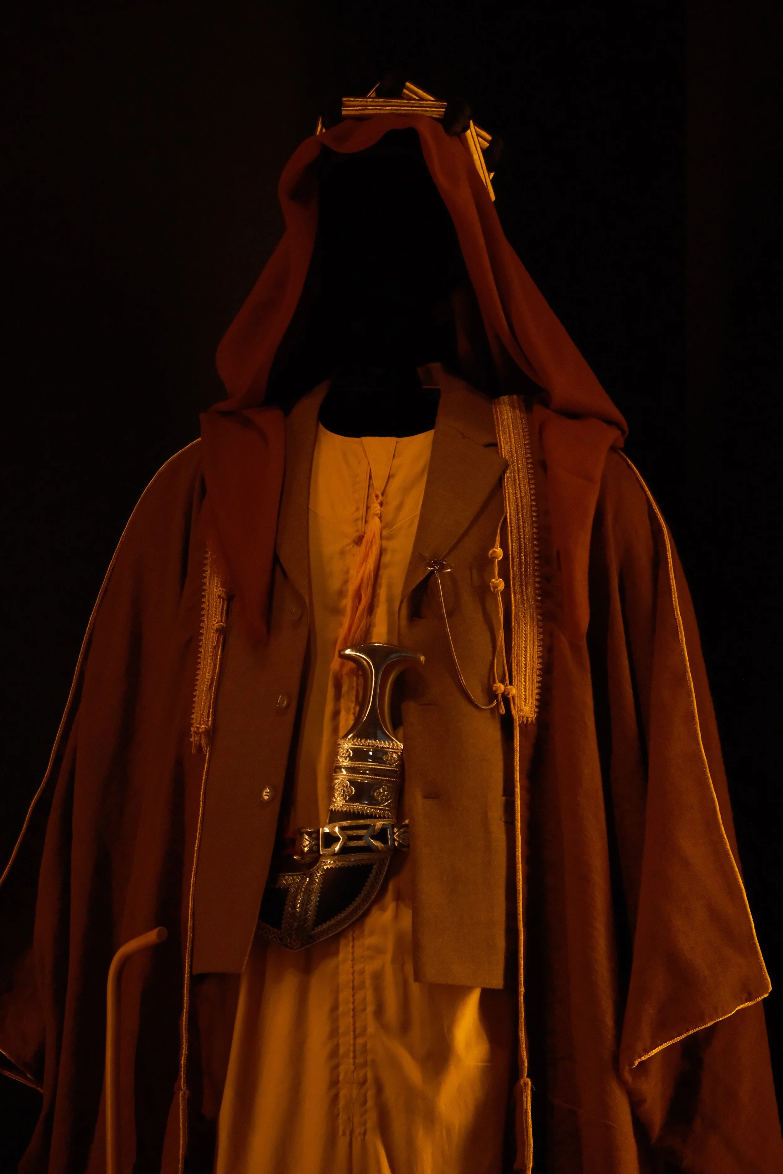 Close-up of a figure wearing a brown cloak with a black middle layer, a belt with a decorative buckle, and holding a silver sword with a detailed hilt, against a black background.