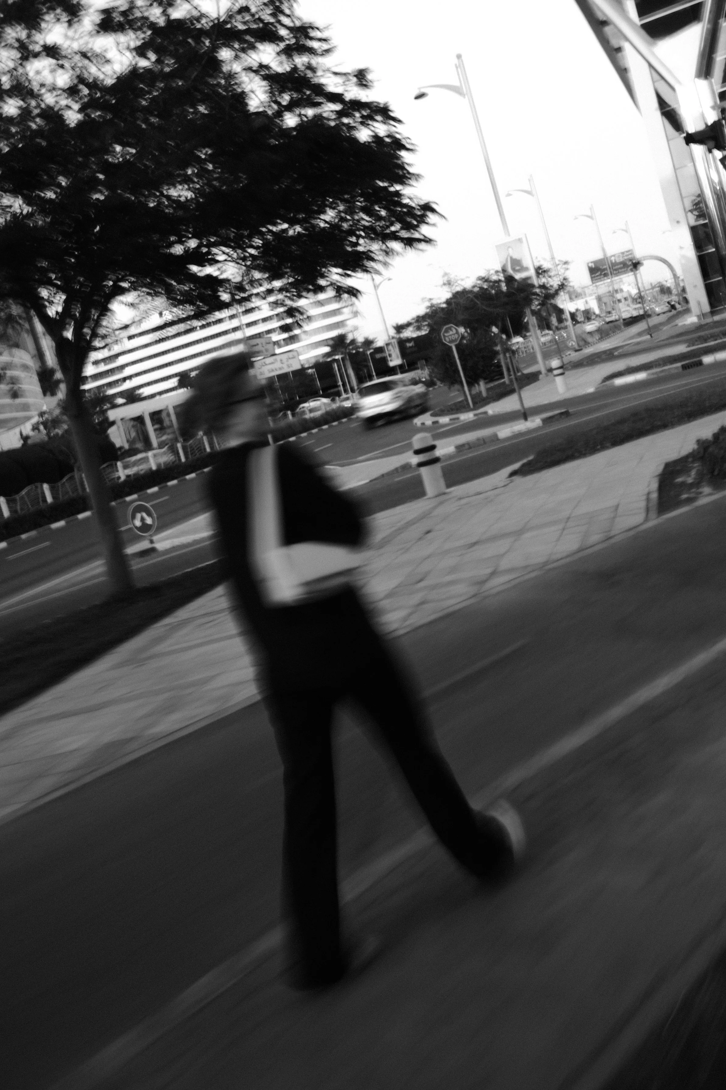 Black and white photo of a city street with a man walking in the foreground, trees, street signs, and buildings in the background.