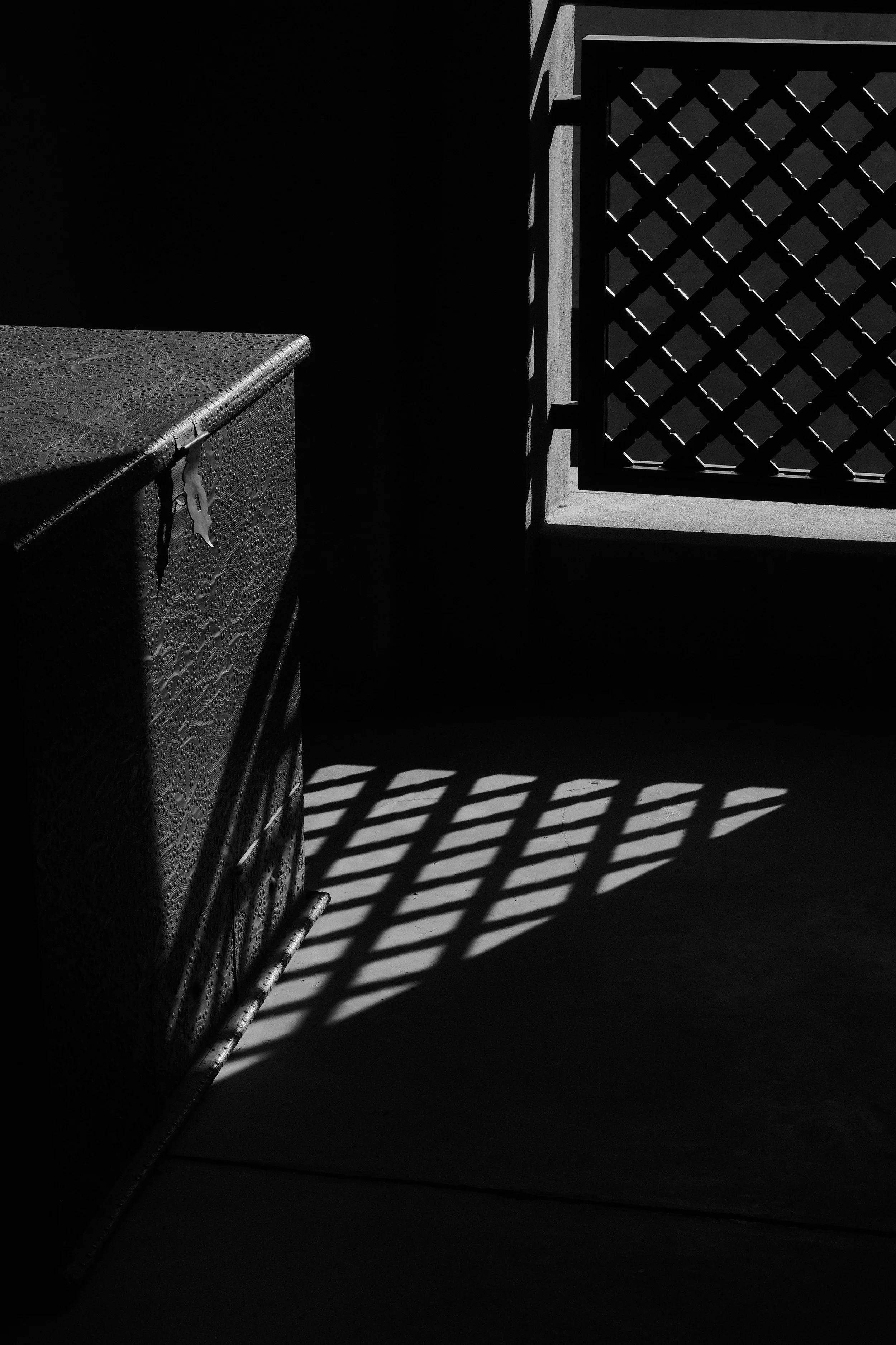 Black and white photo of a textured chest or cabinet on the left and a window with a lattice pattern on the right, casting shadow lines on the floor.