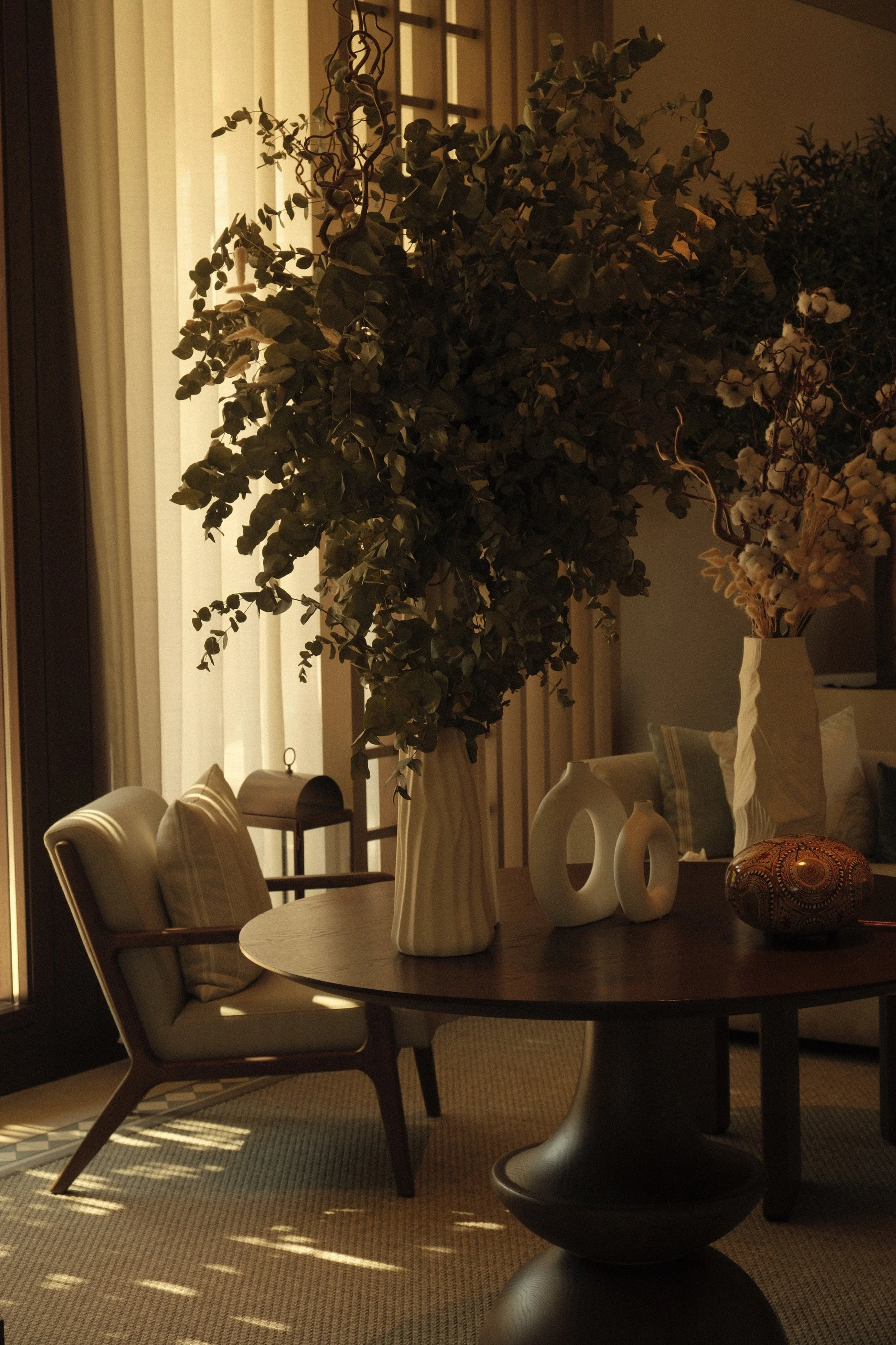 Interior of a dining room with a round wooden table, beige chairs with striped pillows, and decorative vases on the table. There are large green plants near a window with vertical blinds, and sunlight is coming through the window. A small lantern sit