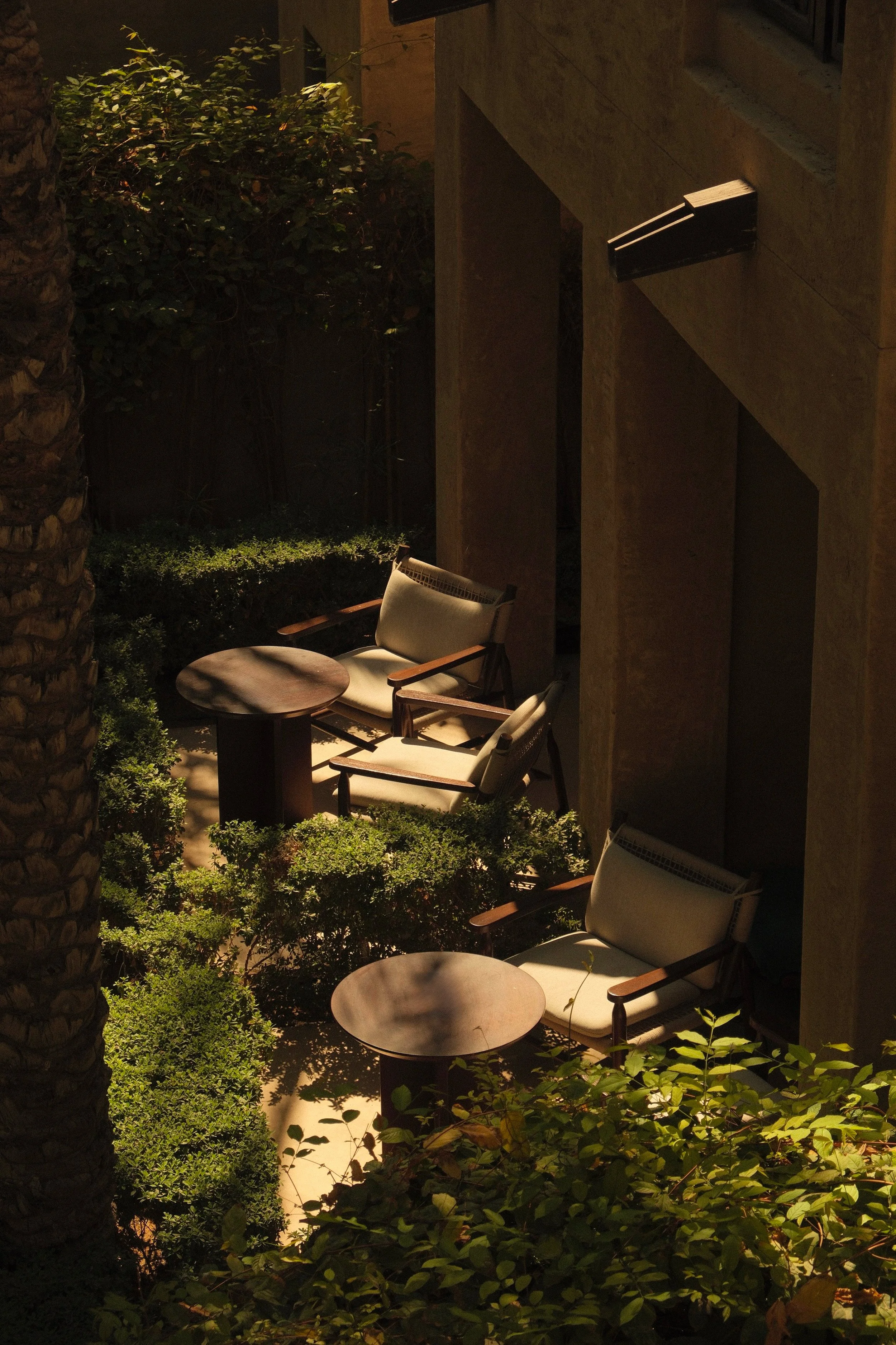 Nighttime outdoor patio with two beige chairs and two small round tables surrounded by lush green bushes and trees, with part of a building and window visible.