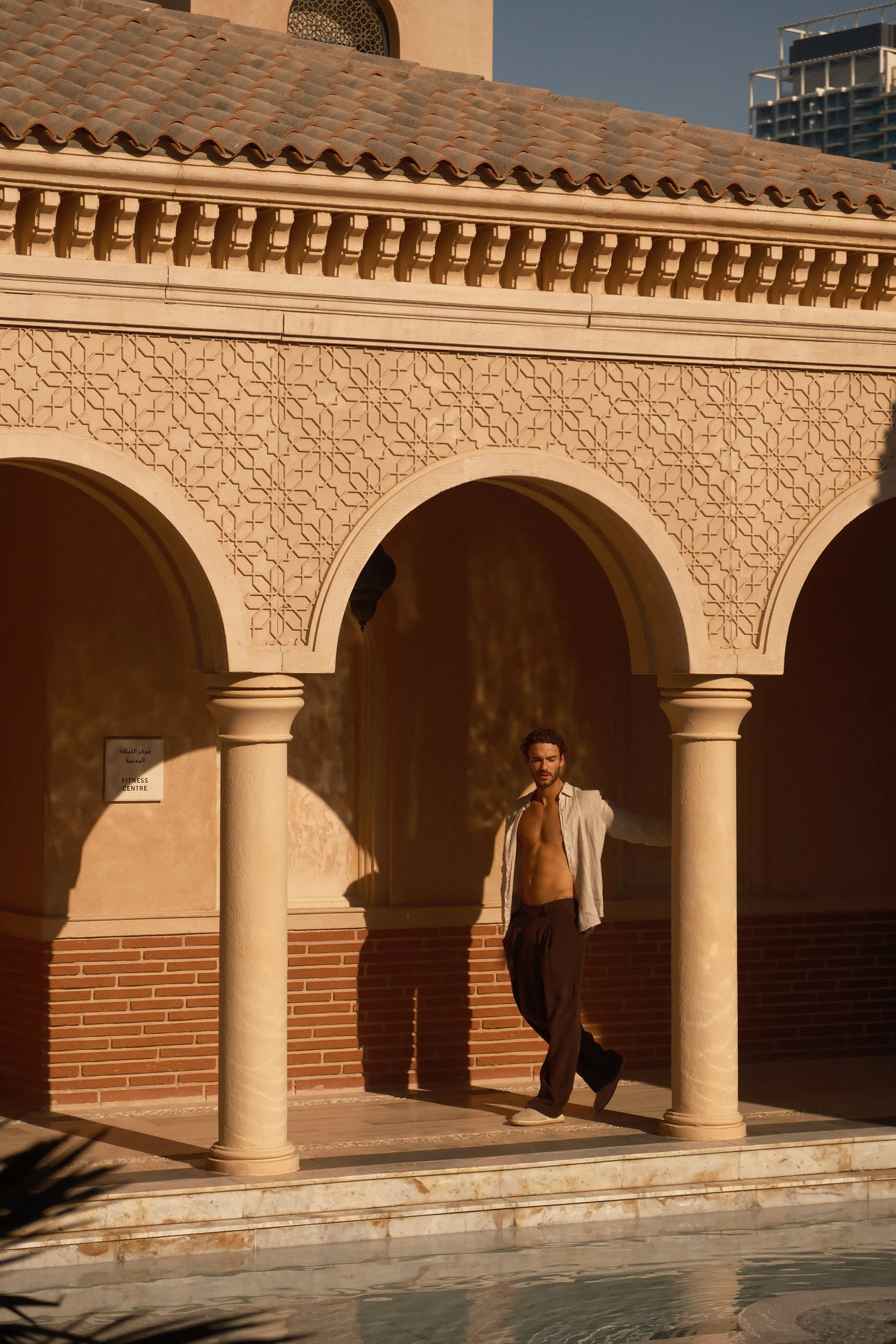 A man with curly hair and a beard is standing under a architectural structure with arches and columns, wearing an open shirt and dark pants, in bright sunlight.