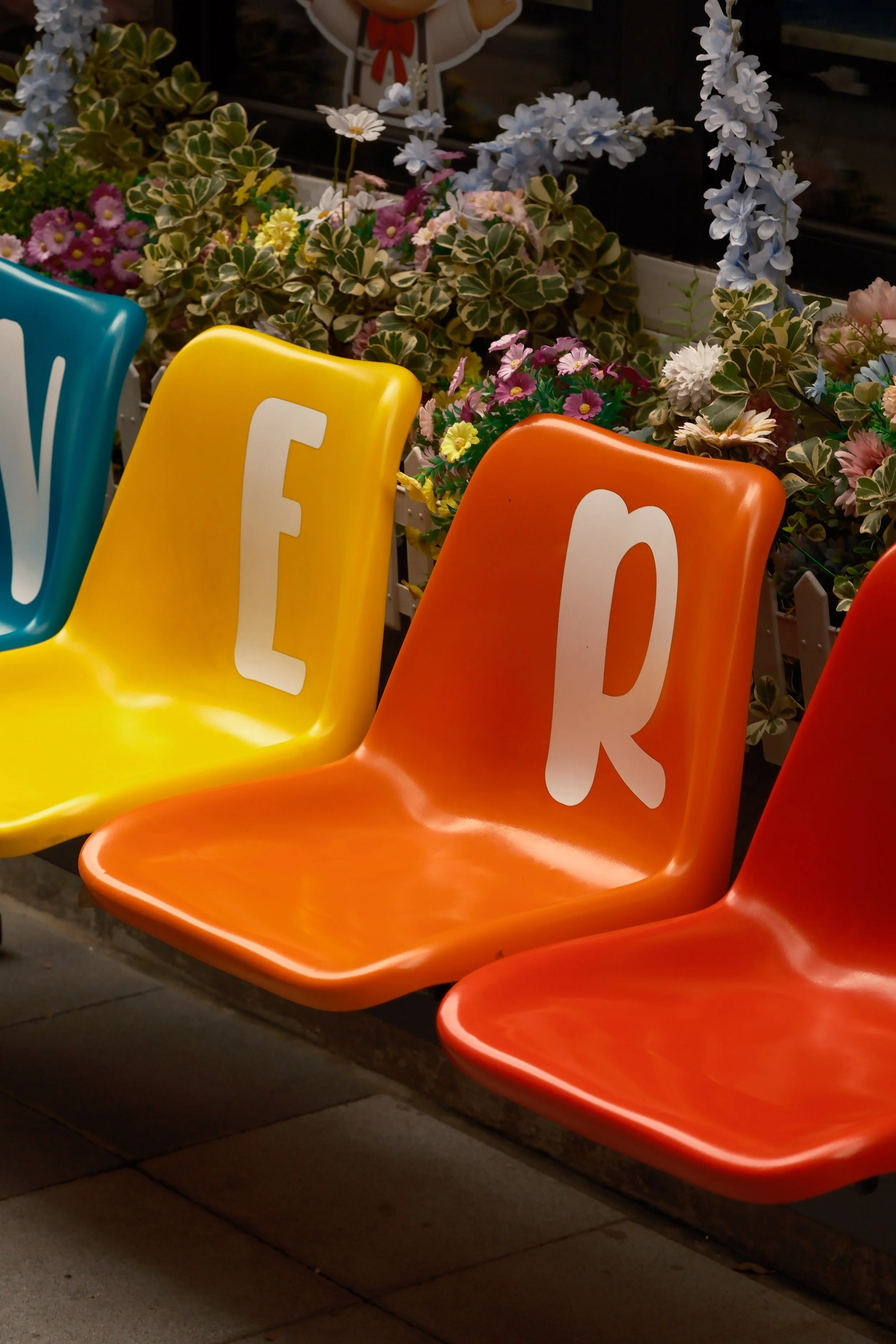 Colorful chairs with white letters, green plants with pink, white, and purple flowers, and a small cartoon bear figure in the background.