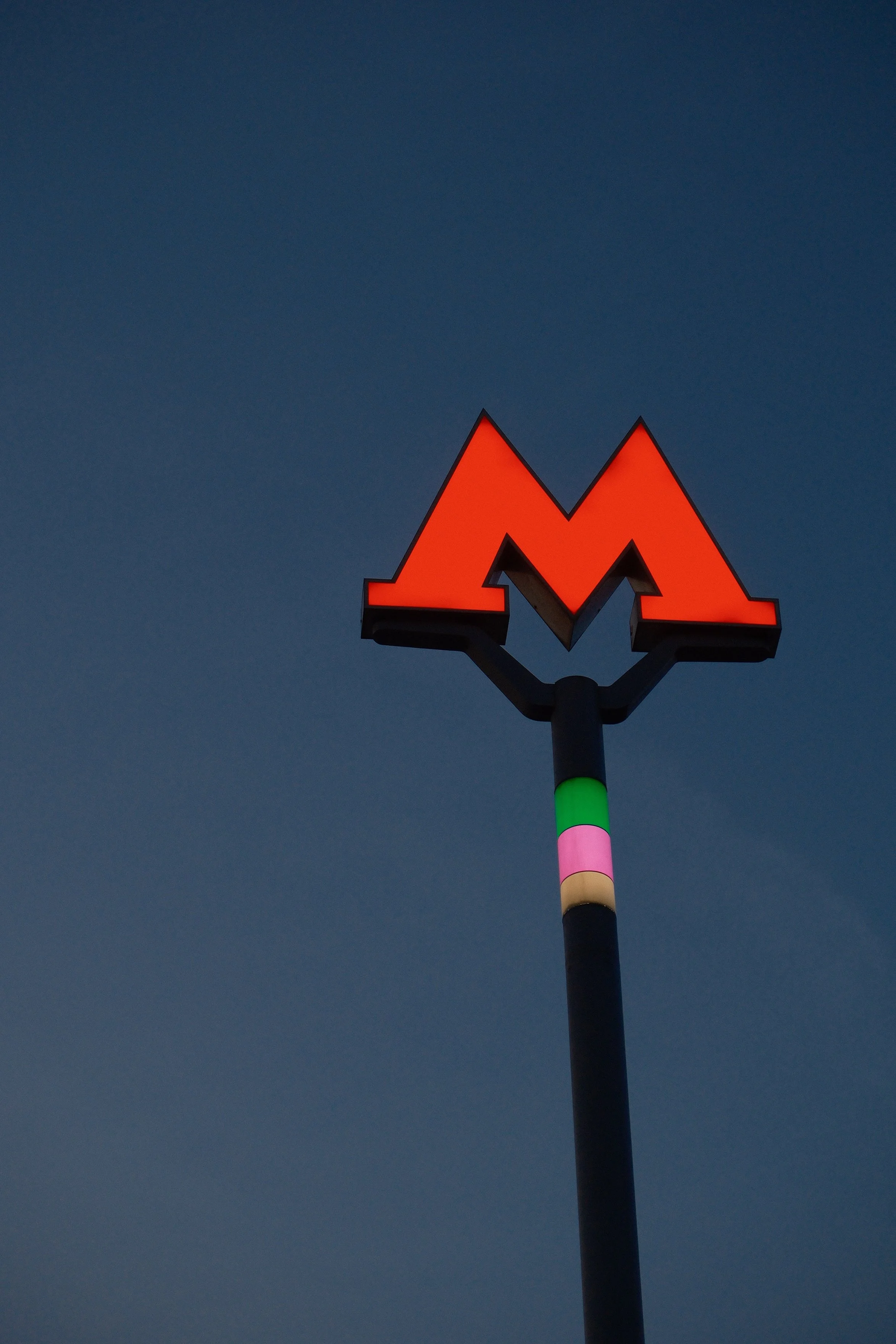 Illuminated orange neon letter M on a tall black pole with colorful sections, set against a dark sky at dusk.