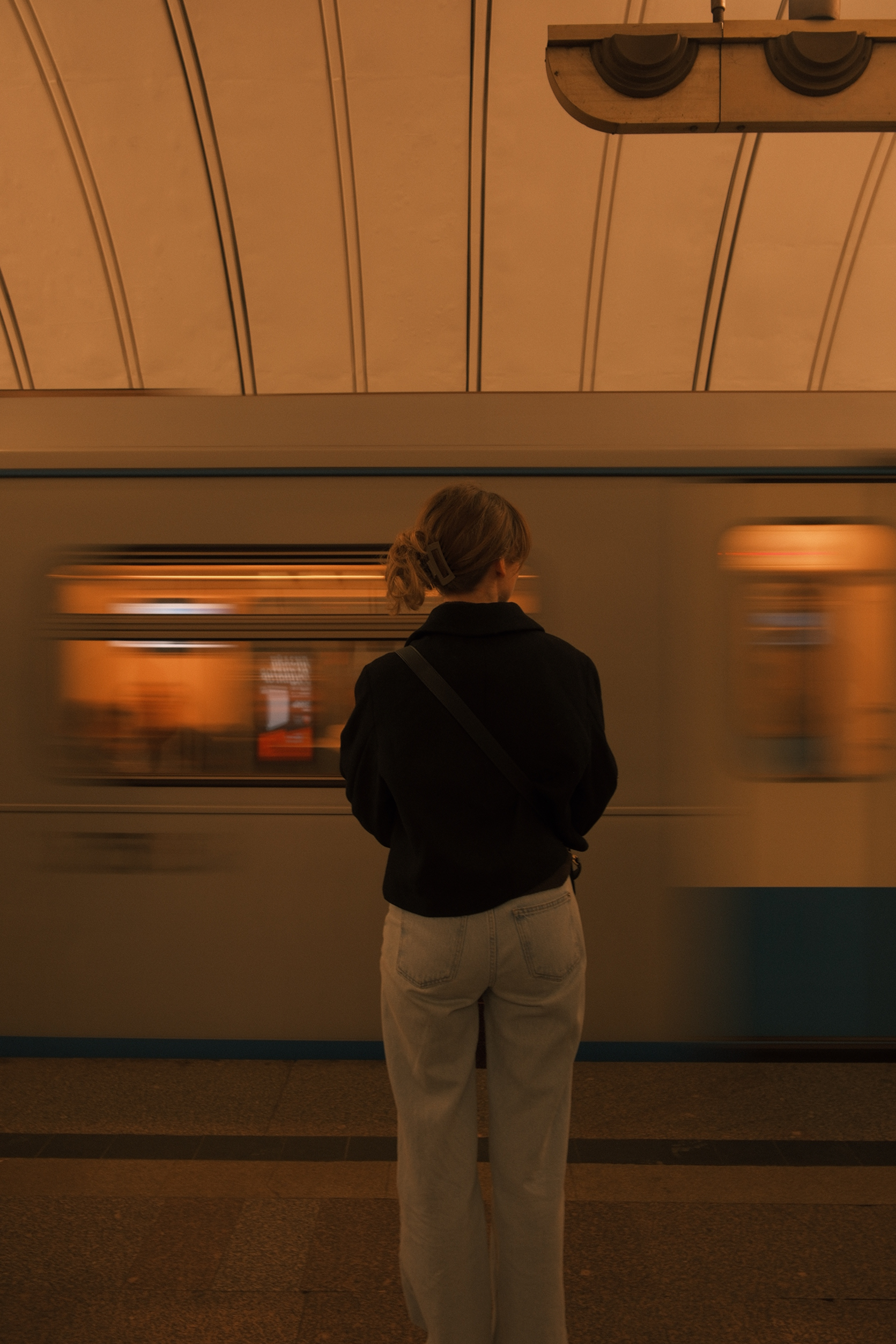 A woman with light brown hair tied back, wearing a black jacket and beige pants, stands in front of a moving subway train.