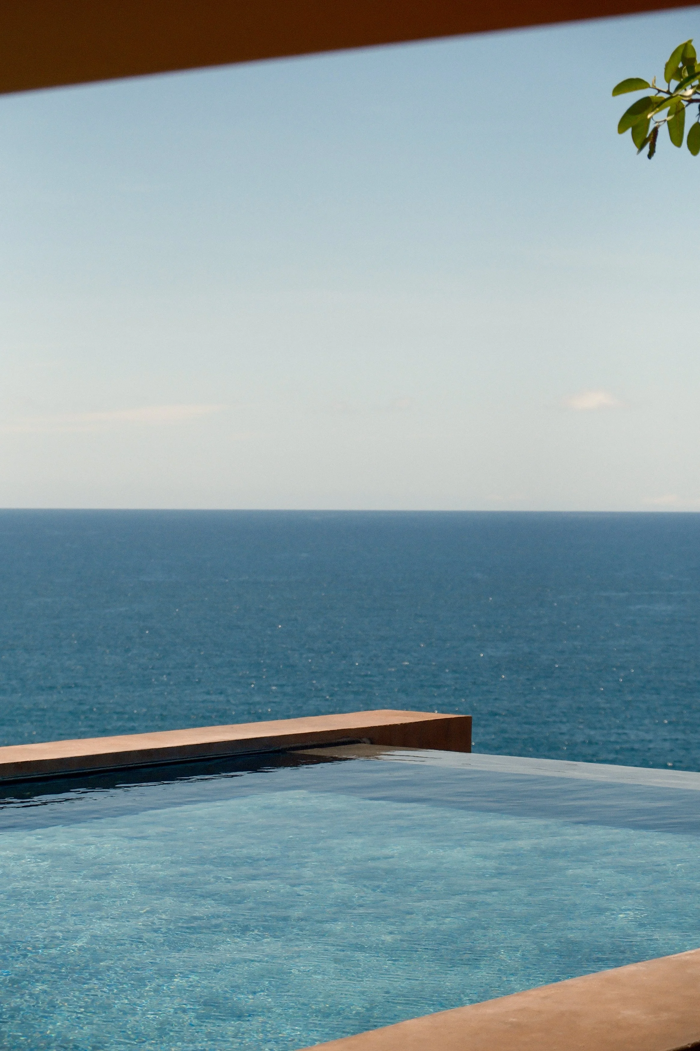 Ocean view from a balcony, with a pool in the foreground, a wooden railing, and a clear sky with some clouds.