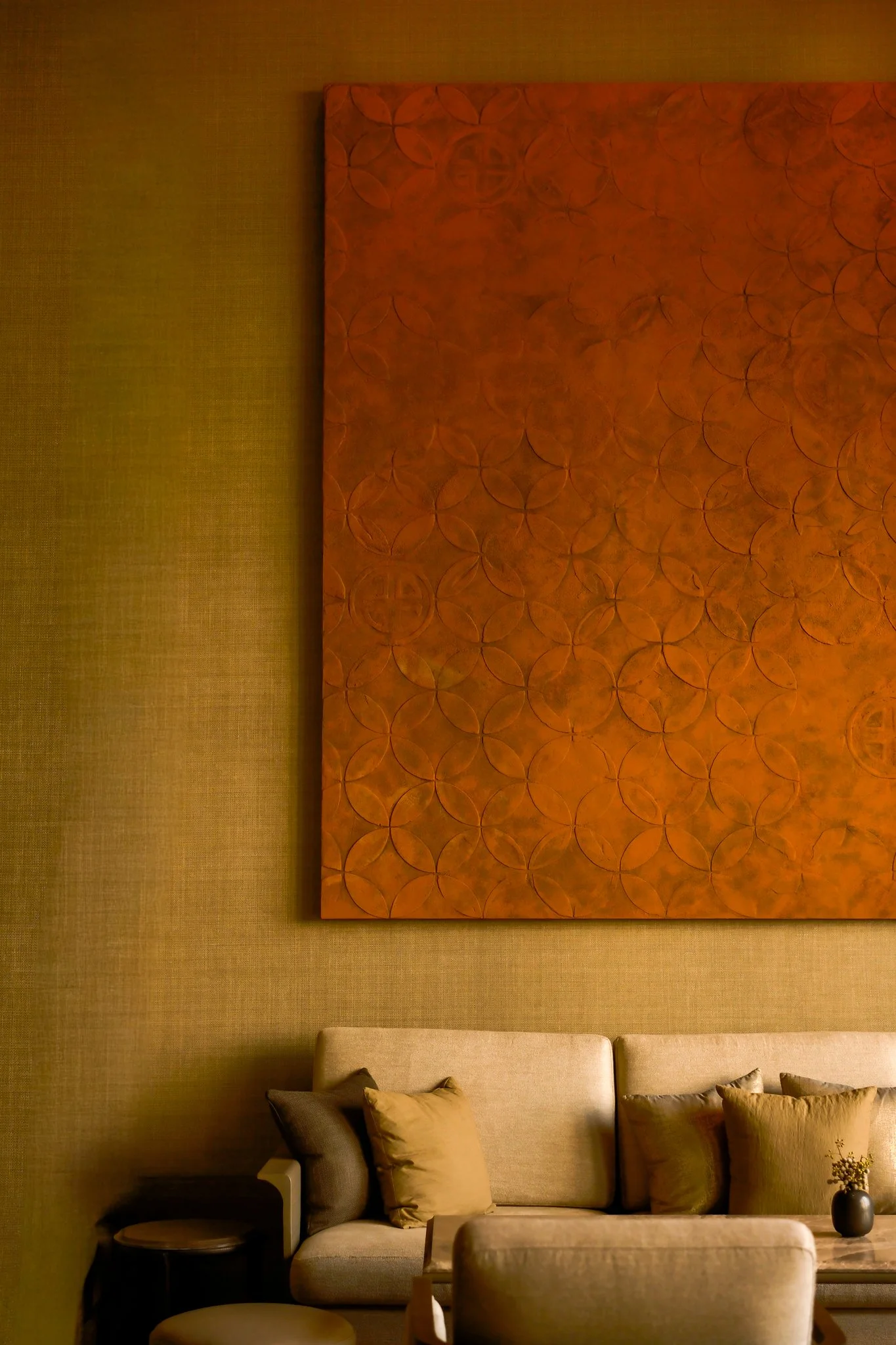Living room with beige sofa, pillows, a side table, a black vase with flowers, a large orange artwork, and textured beige walls.