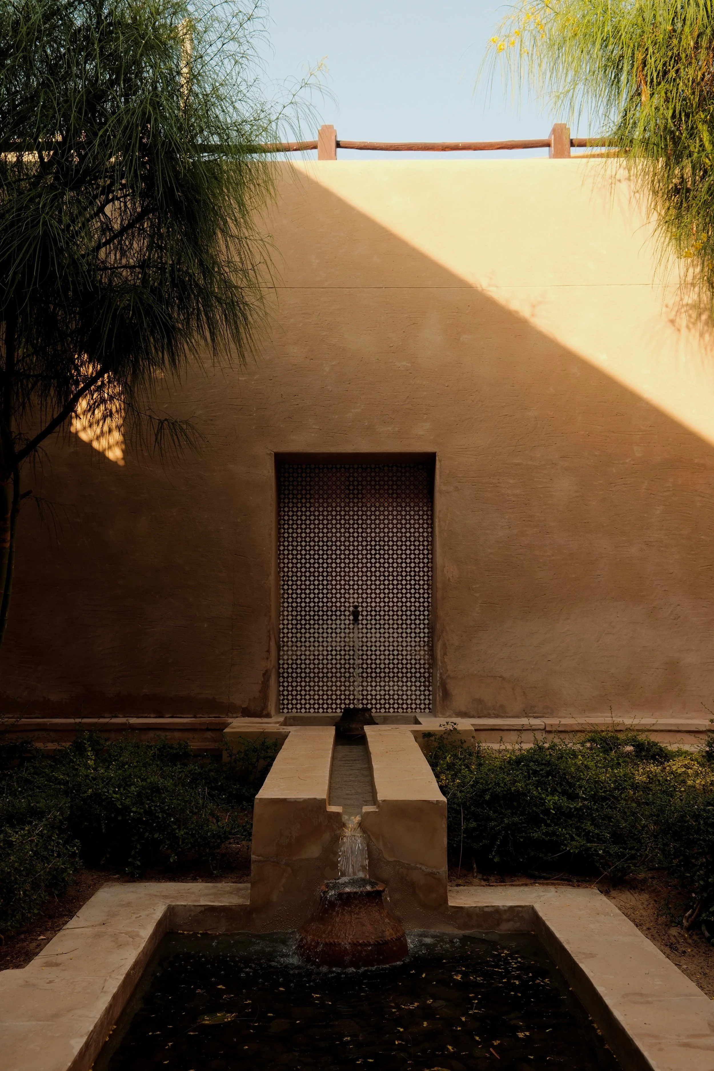 Front view of a modern building with a beige wall, a patterned door, a small fountain in front, and greenery on both sides, under a clear sky.