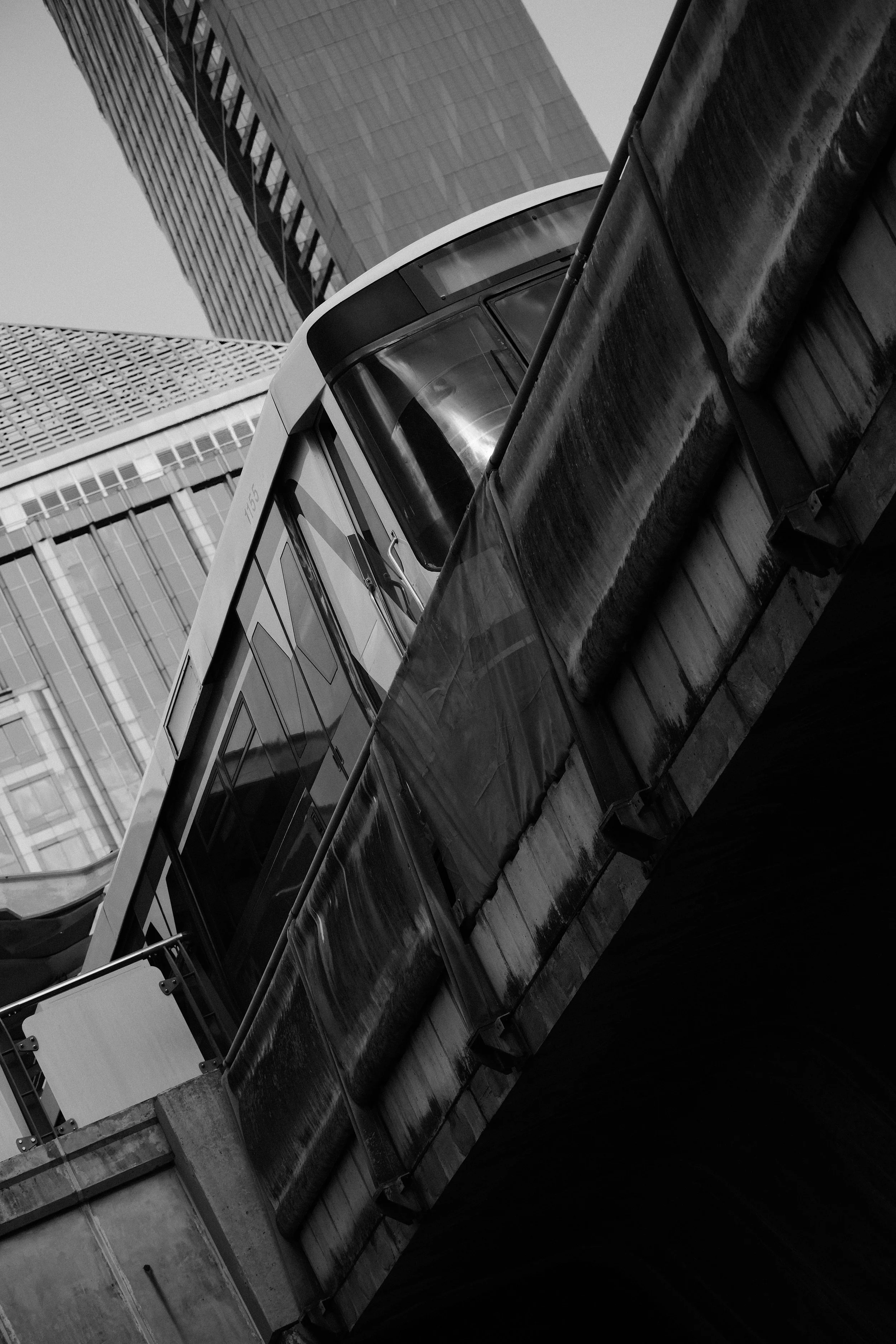 Black and white image showing a train on an elevated track with tall modern buildings in the background.