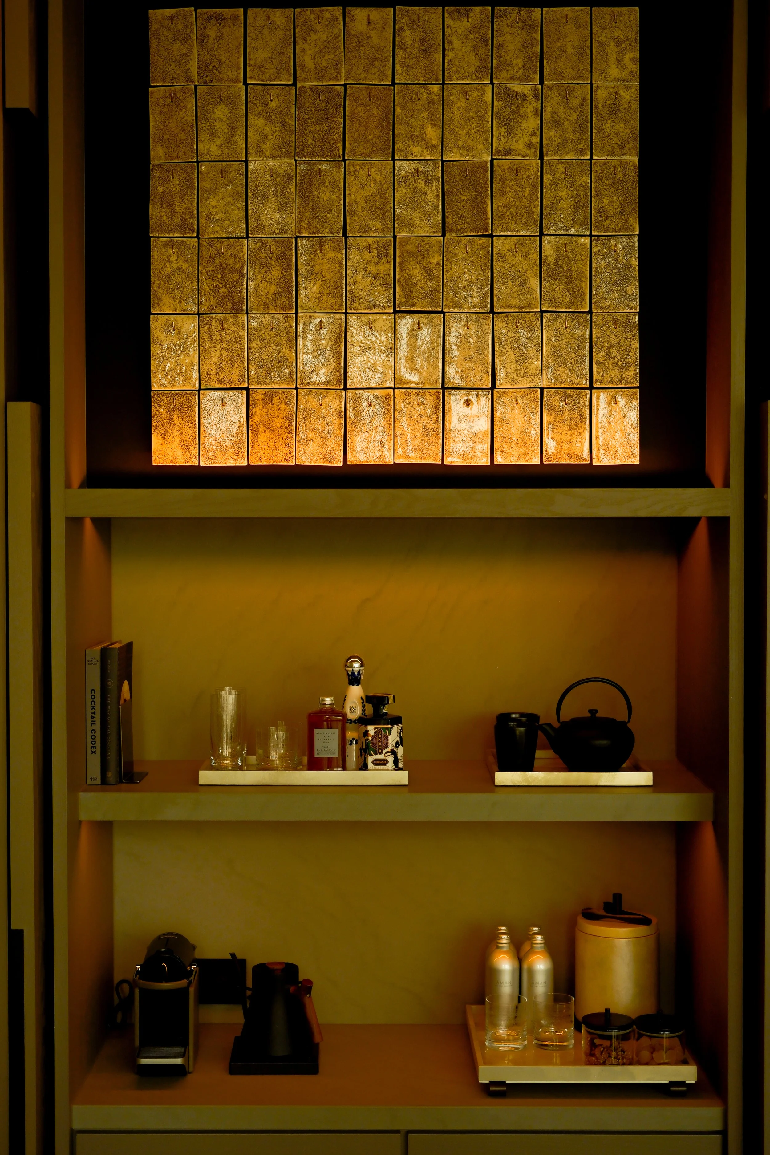 A shelving unit with a backlit textured panel on top, containing various bottles, glasses, a teapot, books, and kitchen appliances.