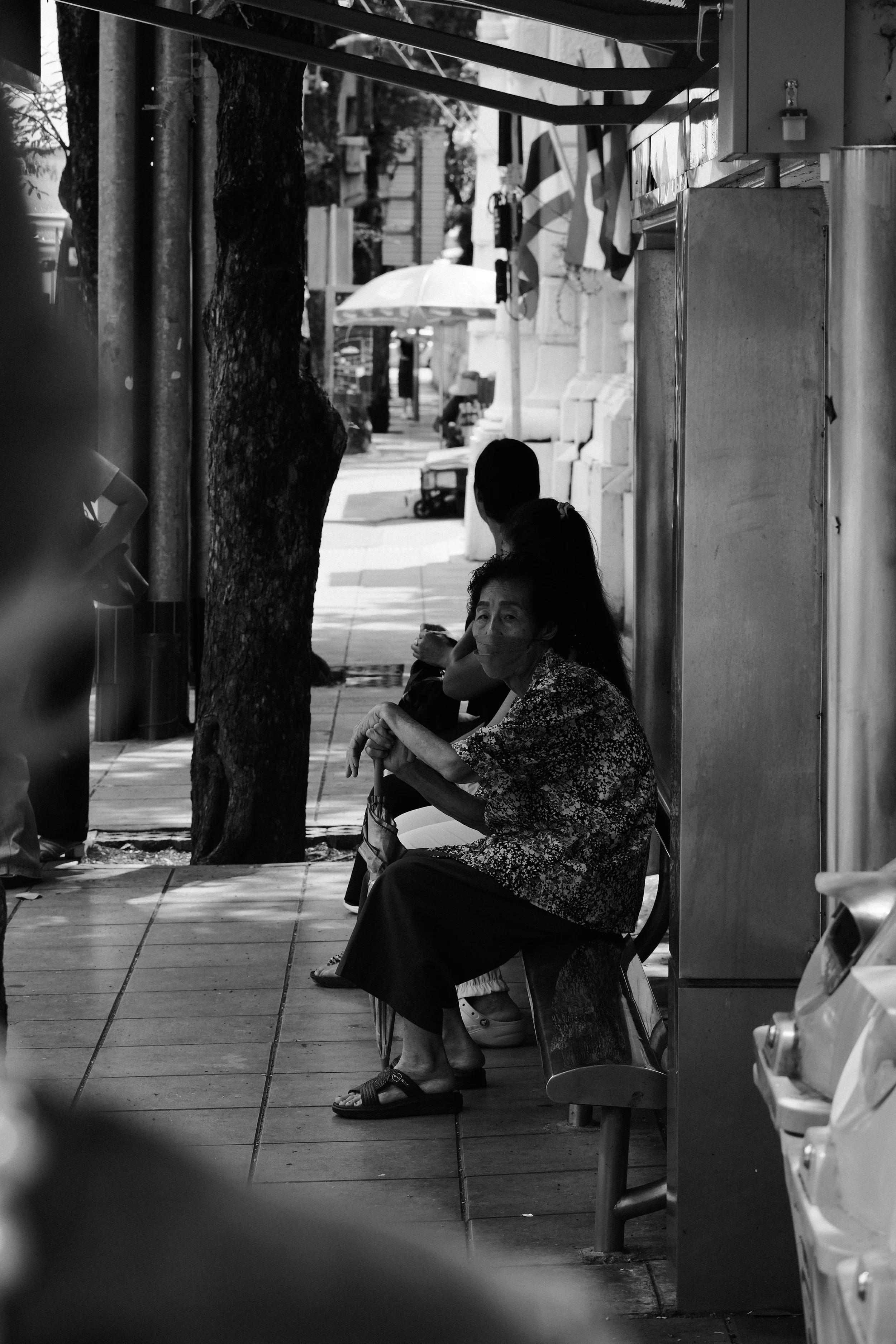 A street scene with multiple women sitting on benches in a sidewalk area. One woman in the foreground is wearing a patterned top and a face mask, looking at the camera. Other women are sitting further away, some facing away. There are trees and build