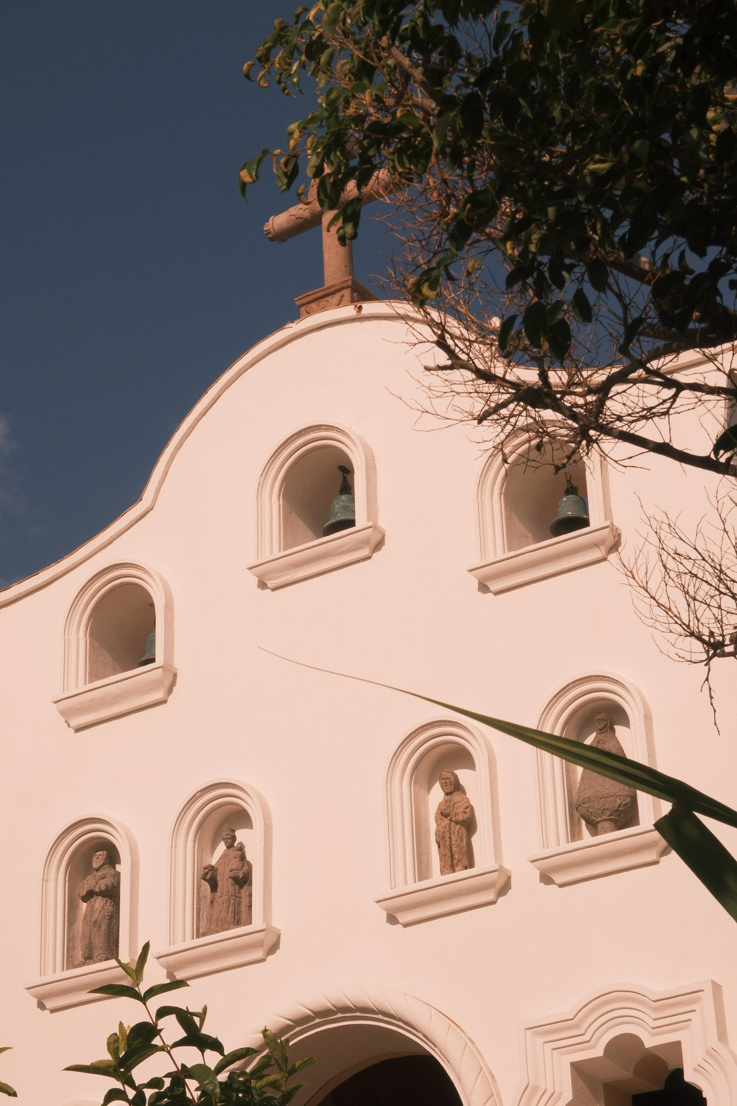 The image shows a white church or chapel with arched windows that each contain a statue or sculpture inside. The roof has a cross on top, and there are trees with green leaves in front of the building, partially obscuring the view. The sky is clear w