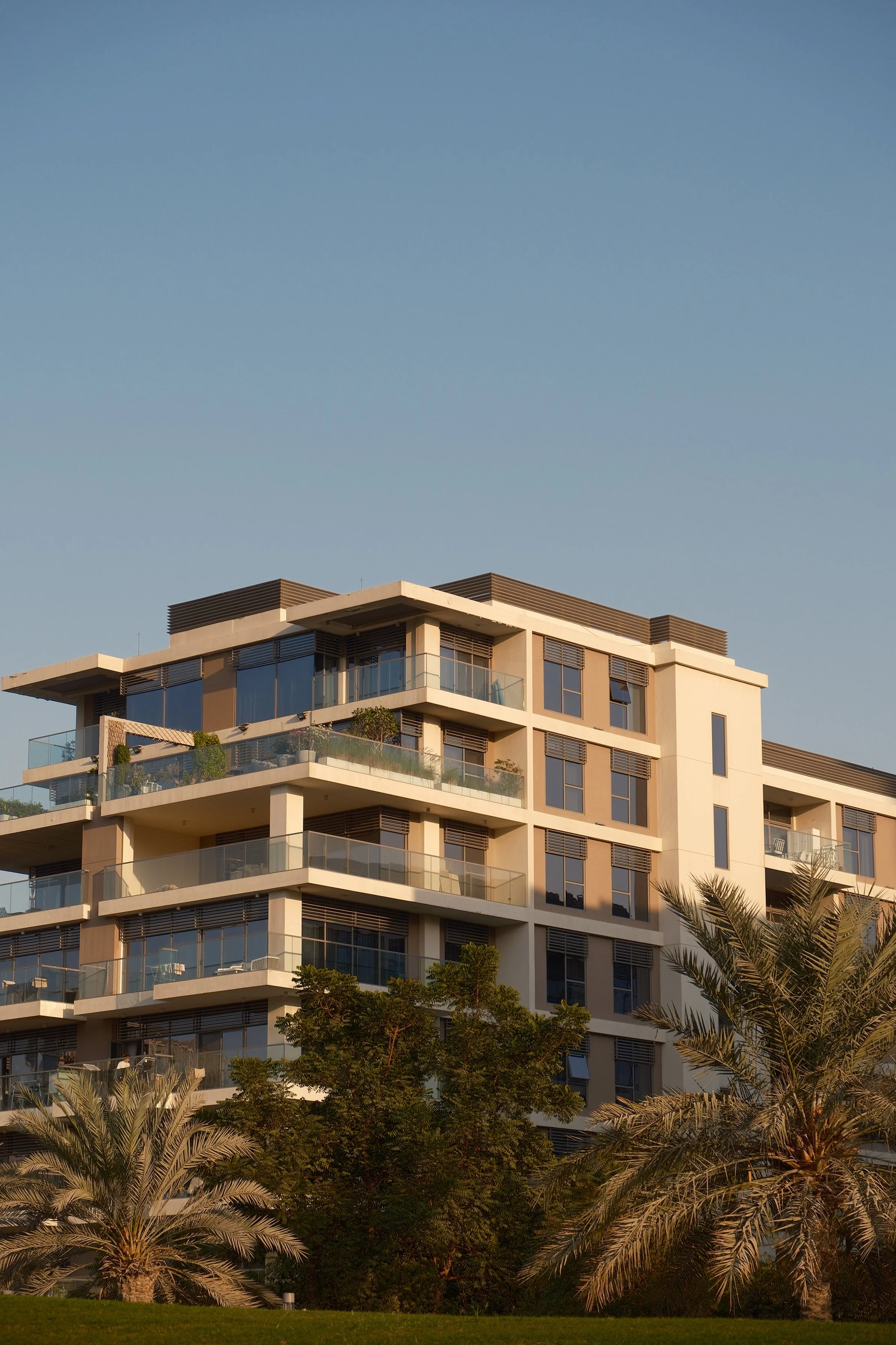 A modern multi-story apartment building with glass balconies, surrounded by palm trees and greenery, under a clear blue sky.