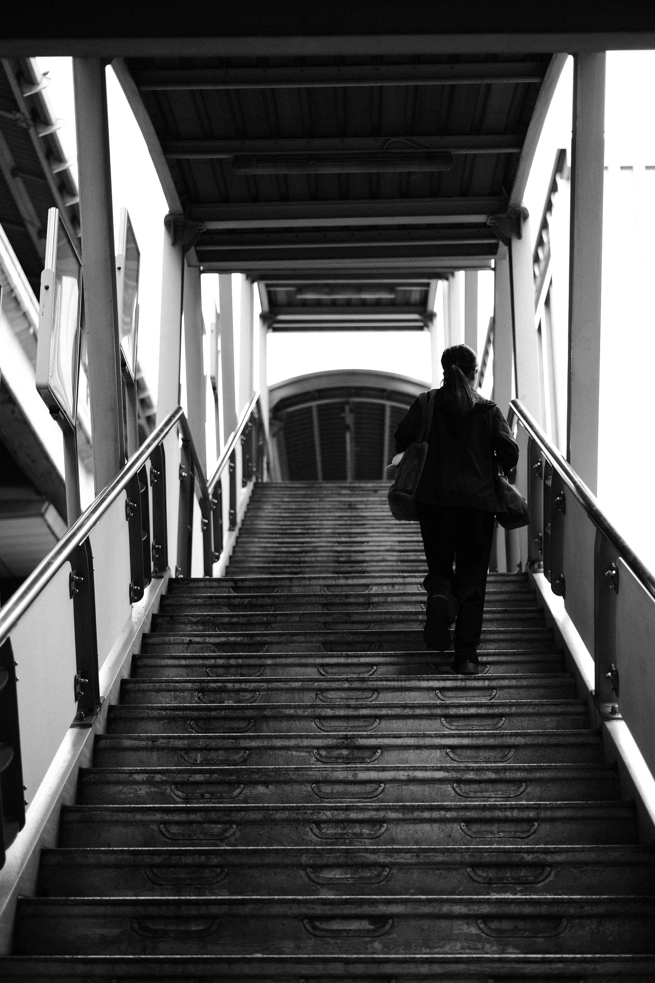 A person with a backpack walking up a metal outdoor staircase at an airport or train station, taken from below and in black and white.